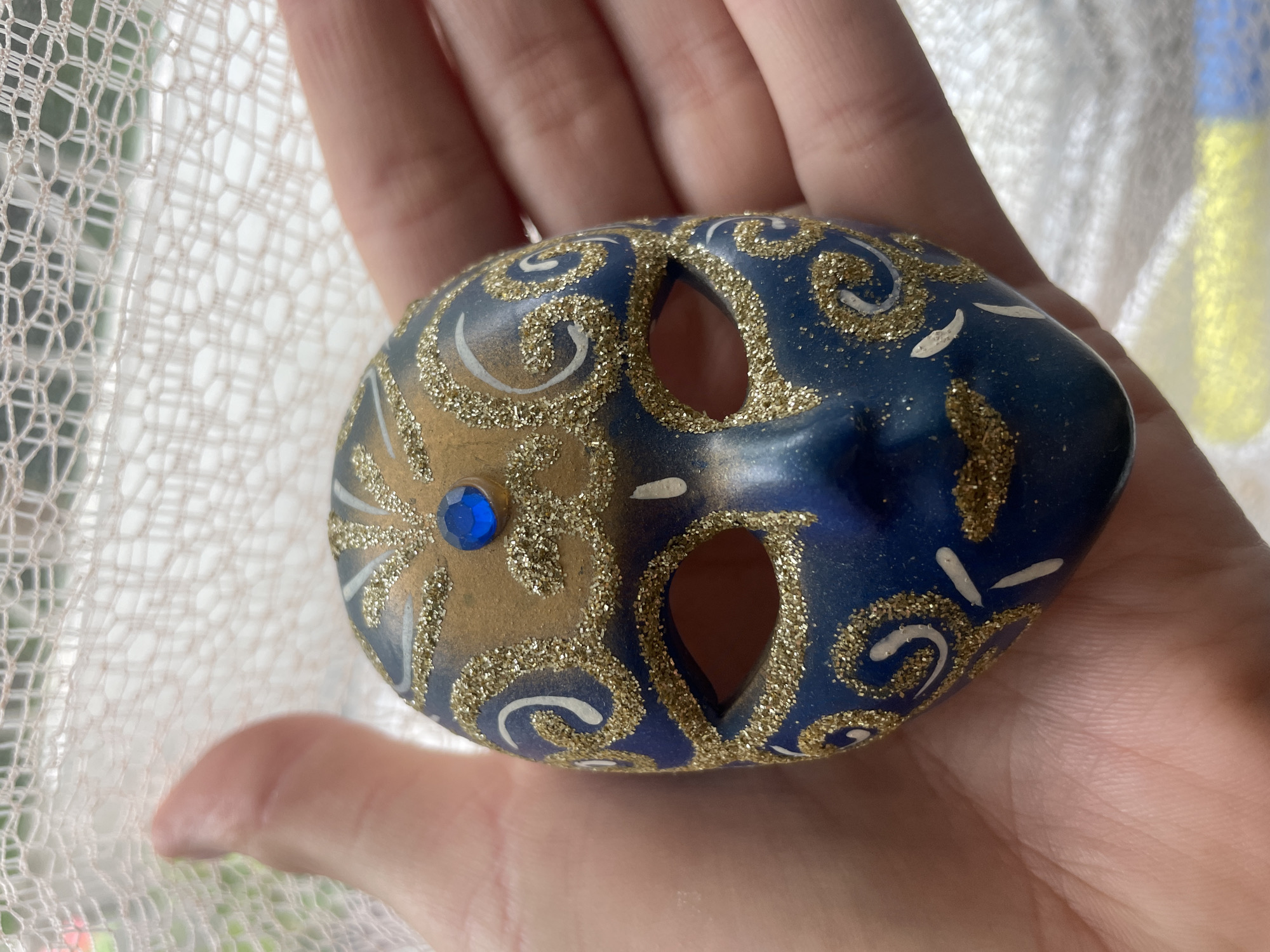 A person holds a small, decorative mask in their hand. The mask is oval-shaped and primarily blue, with gold glitter and white paint accents forming swirling patterns. It has two eye holes and a blue jewel in the center. The person's hand is fair-skinned. The background is slightly blurred but appears to be a window with a white curtain. There is no verifiable evidence in the image to connect this image to the city of Sochi, Russia.