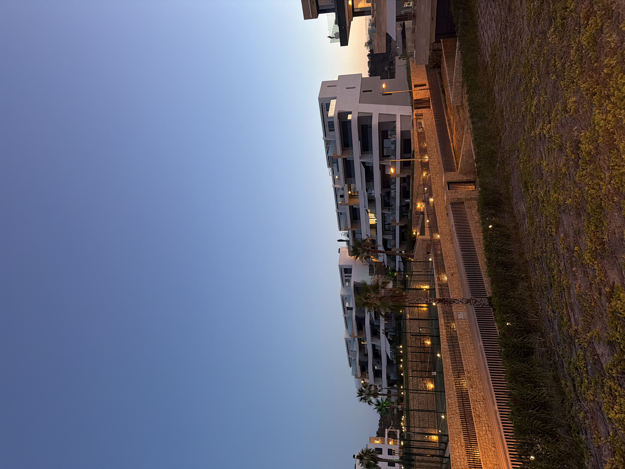 This is a photograph of a modern residential complex in Finestrat, Spain, taken during dusk. The sky is a clear, pale blue, transitioning to a slightly darker shade at the horizon where a faint glow suggests the setting sun. The architecture is characterized by clean lines, white facades, and multiple balconies with glass railings. The buildings are tiered, with the highest structures on the left and right of the frame.
In the foreground, a dry, sandy area is covered with sparse, low-lying green vegetation. Behind this, a pathway lined with stone walls and vertical dark wood or metal slats runs horizontally across the image. Small, warm-toned lights are embedded in the stone walls, illuminating the path and the retaining walls of the structures above. Palm trees are visible, interspersed with the buildings, their silhouettes contrasting against the twilight sky.
A fenced area, possibly a sports court, is situated on a raised level behind the stone walls. It is lined with small, round lights that are illuminated. The overall scene conveys a sense of modern luxury and tranquility, with the artificial lighting creating a warm and inviting atmosphere against the natural fading light of the day. No people are clearly visible in the frame, but the presence of balconies and structures suggests occupancy.