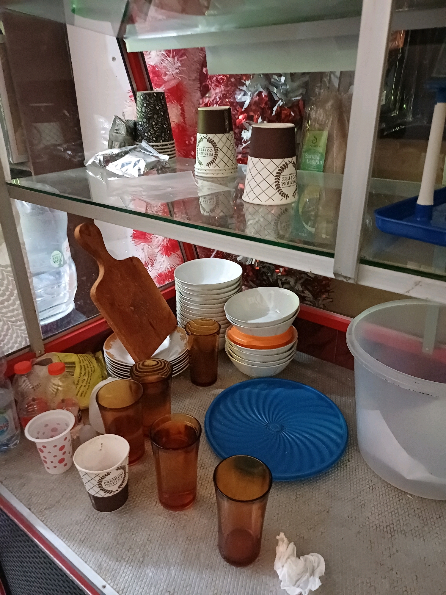 The image depicts a glass display cabinet, likely a food stall or a small shop, located on a street in Dumai City, Indonesia. The cabinet contains various items including stacks of white bowls, orange plates, and brown plastic cups, as well as paper cups with coffee branding. There are also two brown glass cups, a wooden cutting board, and a blue plastic lid on the counter. Bottles of water and other liquids are visible on the left side of the cabinet, along with a bag of snacks. The background of the cabinet shows decorative red and white tinsel, and various packaged goods. The scene appears to be indoors or covered, with a grey textured surface on the counter.