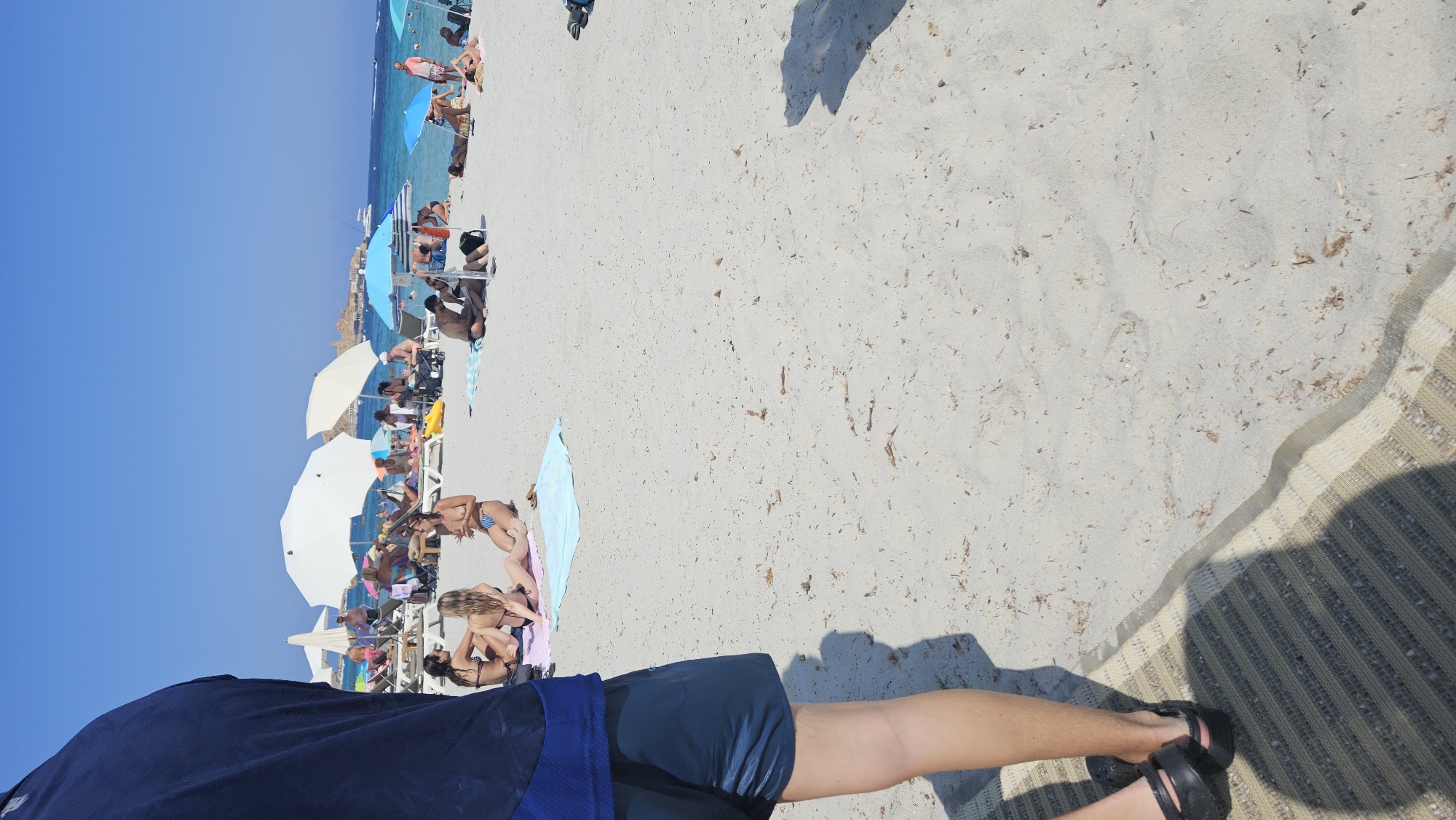 The image captures a bustling beach scene on a sunny day in L'Île-Rousse, France. The foreground features the lower body of a person wearing navy shorts and black sandals, standing on a patterned mat. Beyond this, the sandy beach is populated with numerous people engaged in various activities. Several individuals are sunbathing on loungers or towels, while others are sitting or walking along the shore. The turquoise water is visible in the background, with a few boats on the horizon and a rocky outcrop or island. White and blue beach umbrellas provide shade for some of the beachgoers. The overall atmosphere suggests a lively and enjoyable day at the beach, with clear blue skies overhead.
