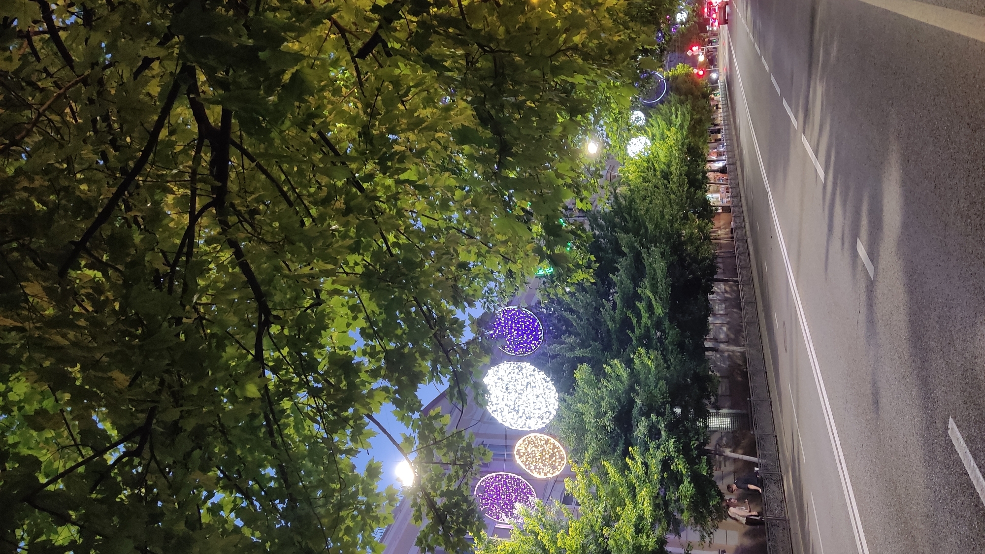 The image depicts a street scene in Krasnodar, Russia, likely at dusk or early evening, as evidenced by the streetlights and decorative lights that are illuminated. The perspective is from beneath a large tree with lush green foliage, which partially obscures the view of the street. The tree's branches and leaves are a prominent element in the foreground.
The street is paved with asphalt and has double white lines marking the lanes. There are no cars visible on the road itself, but a red vehicle can be seen in the distance. Along the side of the road, there is a row of trees, and hanging between them are large, circular decorative lights. These lights are patterned and illuminated with various colors, including white, purple, and yellow. Some are fully lit, while others are partially visible. The overall ambiance suggests a festive or decorative occasion, perhaps for a holiday or event.
In the middle ground, along the sidewalk, a few people are visible. One person is leaning against a fence, and another pair appears to be conversing. A building with windows can be seen behind the trees and lights. The lighting is dim, creating shadows and highlighting the illuminated decorations. The weather appears to be clear, with a visible blue sky peeking through the tree leaves. There is no visible text in the image.
