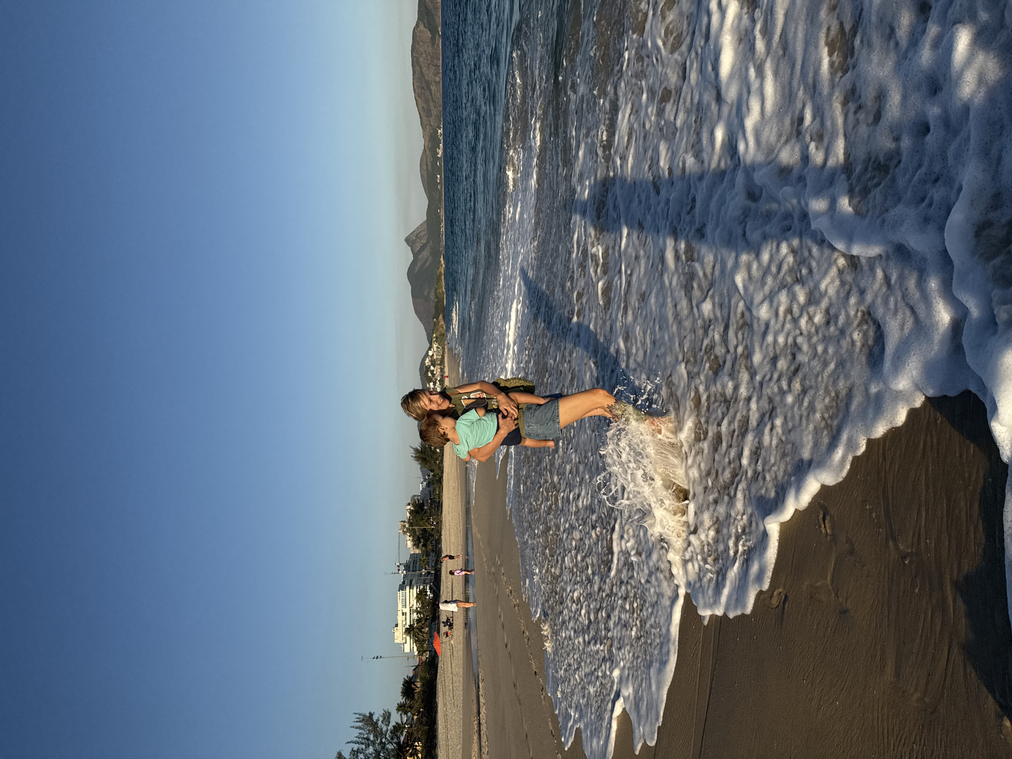 This media file depicts a beach scene in Niterói, Brazil, likely during the late afternoon or early evening due to the long shadows cast by the sun. The sky is a clear, bright blue, transitioning to a lighter hue near the horizon, suggesting pleasant weather. The ocean is calm, with gentle waves washing ashore, creating a foamy edge on the dark, wet sand.
The primary subjects are two people, an adult and a child, standing in the shallow water. The adult is holding the child, and both appear to be relaxed and enjoying the moment. The child is wearing a blue t-shirt, and the adult is wearing a green t-shirt and denim shorts. Their interaction suggests a heartwarming family moment.
In the background, a picturesque coastline is visible, with mountains rising behind a strip of buildings and palm trees. The architecture of the buildings suggests a populated coastal town. Further down the beach, other individuals can be seen, some walking and others seated, enjoying the beach.
The overall mood of the image is serene and joyful, capturing the essence of a relaxed day at the beach. The natural beauty of the location, combined with the simple human interaction, creates a peaceful and inviting atmosphere. There is no visible text in the image.