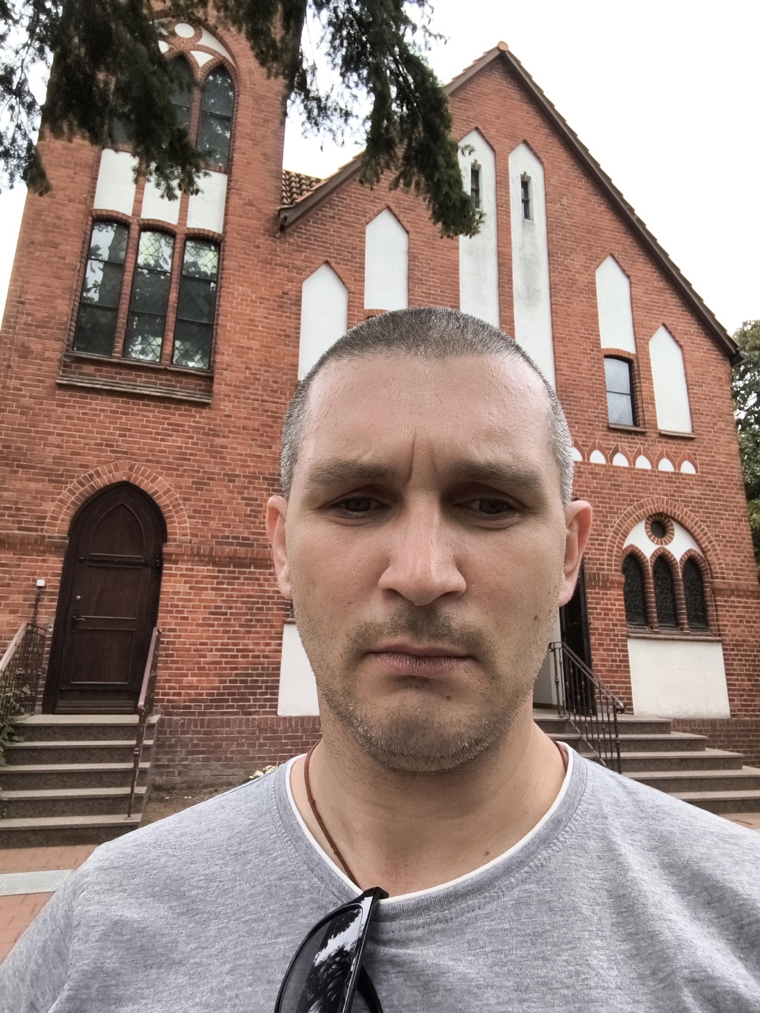 In a close-up, low-angle selfie, a light-skinned male with short, graying hair and stubble is centered in the foreground, looking directly at the camera with a neutral expression. He is wearing a gray t-shirt and sunglasses are visible hanging from his shirt. Behind him, a red brick building with white trim and arched windows is visible. The building appears to be a church or a similar public institution. Trees with green foliage are present in the upper portion of the image, obscuring the sky. The scene is set outdoors, likely in a temperate climate. The building's architecture suggests it is located in a European city, potentially in Germany given the context. This image was taken on the grounds of the Evangelische Kirchengemeinde St. Petri, a church in Wolgast, Germany.