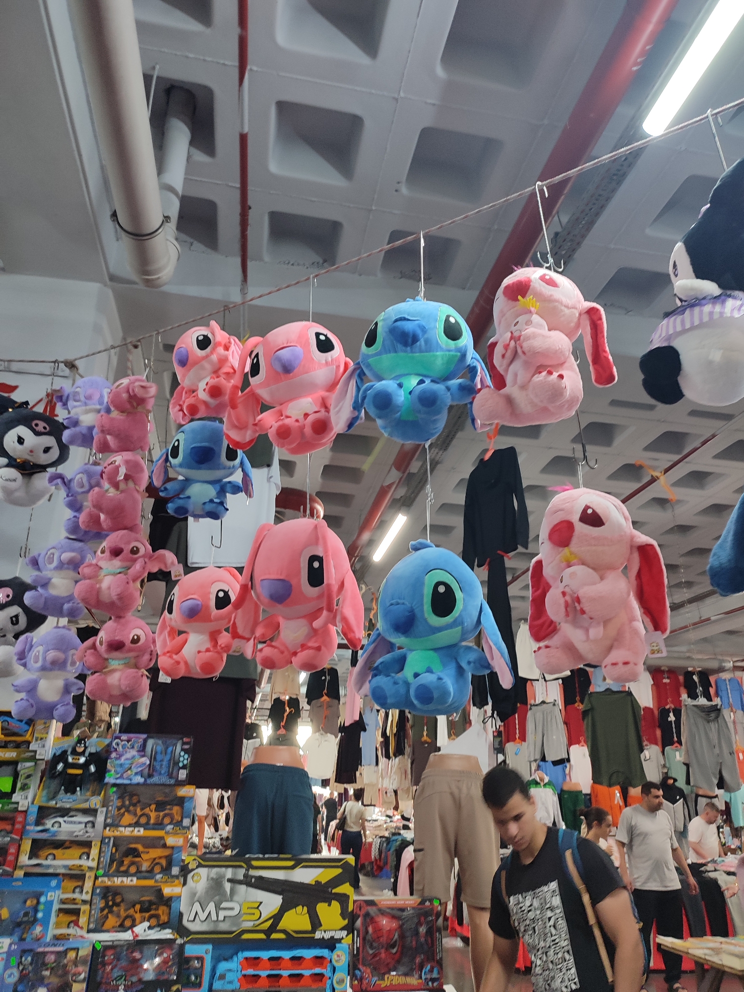 This is a ground-level, eye-level, medium shot of a market stall in Kadıköy, Turkey, featuring a display of plush toys and various clothing items. The plush toys, primarily Stitch and other characters from Disney's Lilo & Stitch, are suspended from the ceiling by strings, creating a vibrant, colorful canopy. Below the toys, a variety of clothing is displayed on racks, including t-shirts, shorts, and jackets, with some mannequins dressed in casual wear. Several people are visible in the background, browsing the merchandise. The market appears to be indoors, with exposed pipes and ceiling tiles indicating a functional, perhaps slightly aged, structure. The overall impression is one of a bustling, commercial environment where toys and apparel are sold.