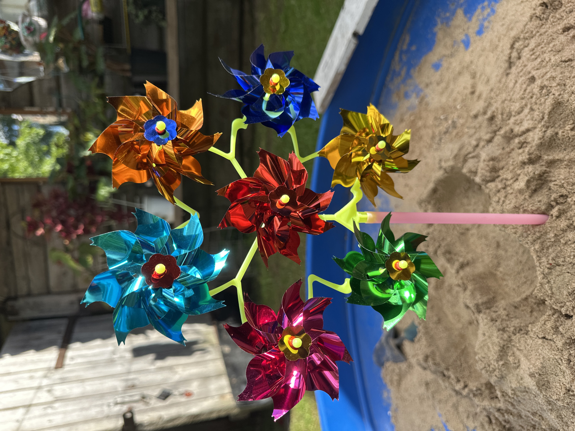 The image displays a colorful pinwheel with seven individual pinwheel flowers. The pinwheels are attached to a lime green stem, which is inserted into a pink tube, presumably a handle. The pinwheels themselves are made of metallic material and come in various vibrant colors: orange, blue, red, gold, light blue, green, and a deep reddish-pink. Each pinwheel has a small yellow and gold center.
The pinwheel is positioned outdoors, with a background that suggests a backyard or garden setting. To the left, there's a wooden fence and some greenery, possibly plants or trees, with some out-of-focus elements that might be garden decorations. The right side of the image shows a blue container filled with sand, suggesting it might be a sandbox or a part of an outdoor play area. The sunlight indicates it is daytime, and the shadows cast by the pinwheel suggest a bright, sunny day.
There is no visible text in the image. The overall mood is cheerful and playful, evoking a sense of summer or outdoor activity. The composition focuses on the pinwheel, showcasing its bright colors and intricate design against the sandy backdrop. The location context of Piriutino, Russia, is provided but not visually evident from the image itself.