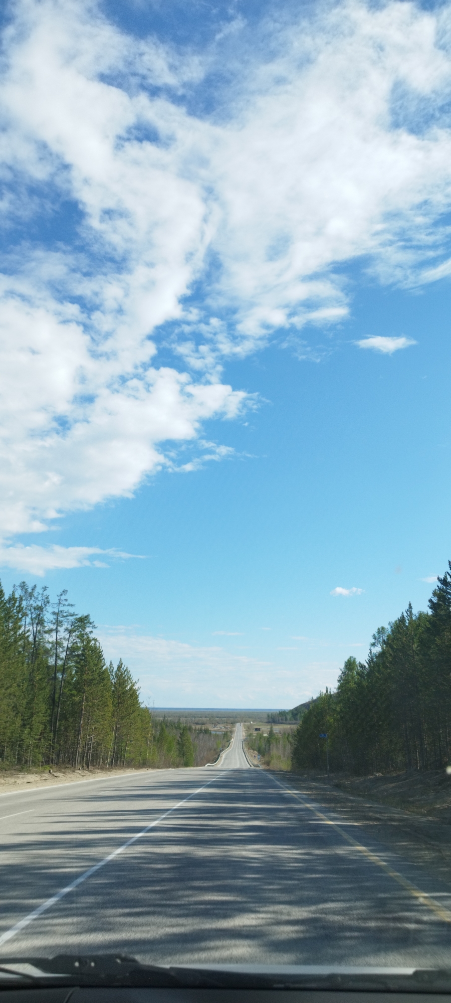 The image depicts a long, straight two-lane road cutting through a forest. The location is Tekhtiur, Russia. Tall pine trees line both sides of the road. The road surface is grey asphalt with a white center line and a yellow line on the right shoulder. Shadowy patterns are cast across the road surface, likely from the trees. In the distance, the road appears to rise slightly over a hill. Beyond the trees, a flat expanse is visible on the horizon. The sky is predominantly blue, with patches of white clouds concentrated in the upper portion of the frame. The image is taken from within a vehicle, with a portion of the dashboard visible at the bottom.