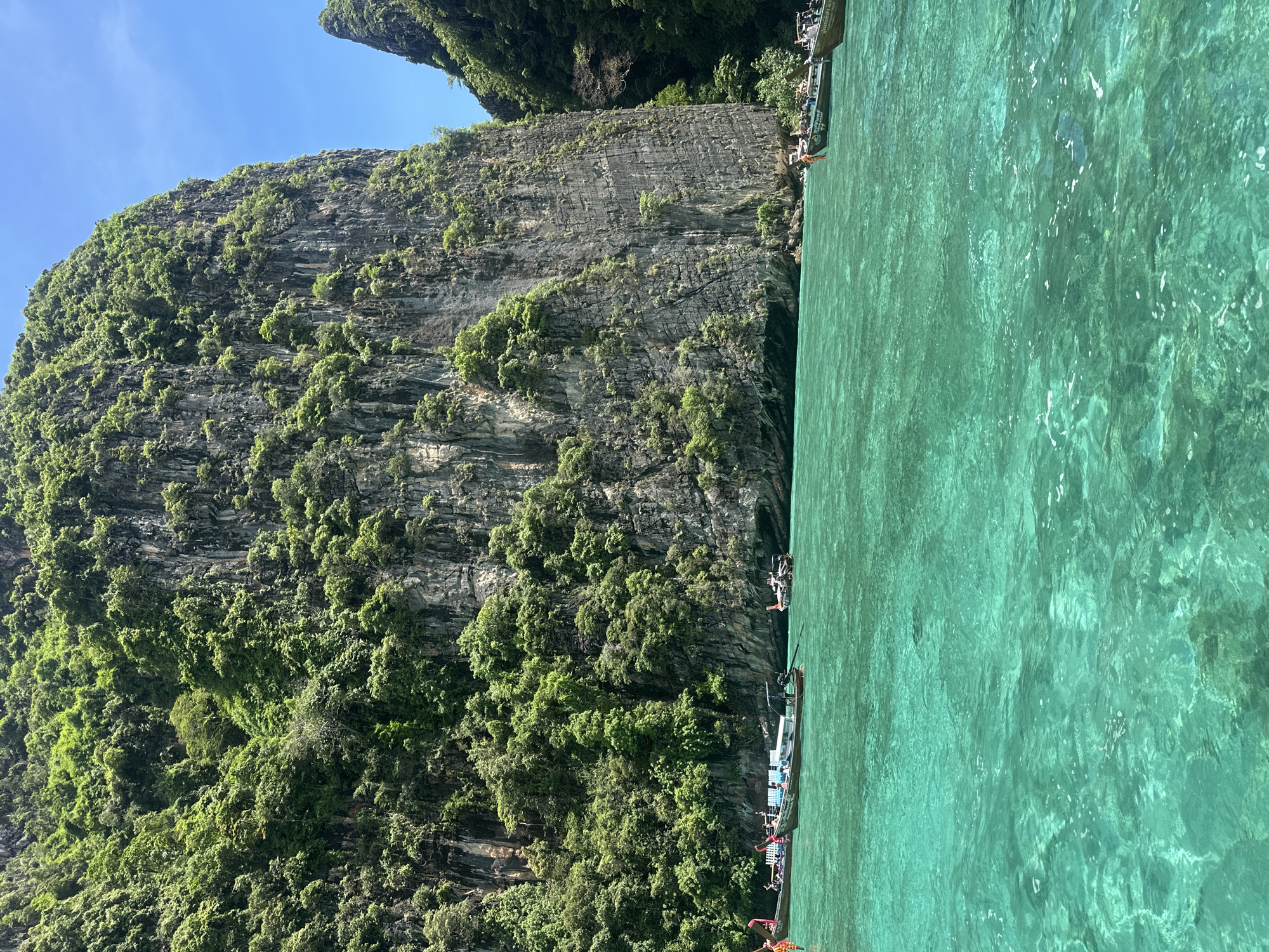 An eye-level shot captures a vibrant turquoise sea against a towering, lush green karst cliff face under a bright blue sky. Several long-tail boats, with their characteristic wooden hulls and overhead awnings, are moored in the clear water near the base of the cliff. The boats are populated with visible passengers, some standing and others seated. The sheer cliff face is densely covered with tropical vegetation, with rugged rock formations visible beneath the greenery. The water's transparency allows the seabed to be faintly discerned in some areas, and small ripples indicate gentle movement on the surface. The scene evokes a tropical island destination, likely in Southeast Asia, given the style of the boats and the geological formations. The location is unknown but is likely a coastal area with limestone cliffs.