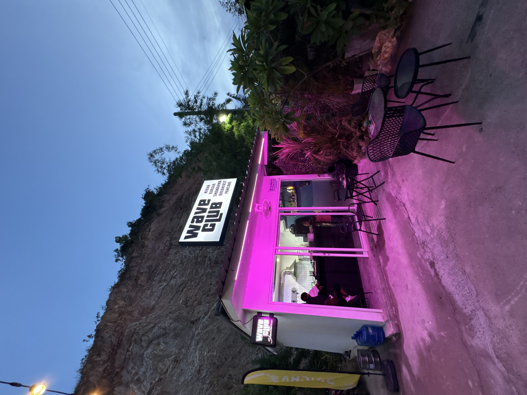The image depicts the exterior of "Wave Club," a food establishment in Baan Bang Rak, Thailand. The building is situated at the base of a rock cliff. A sign displaying "Wave Club" and a list of food items (kebab, burger, shawarma, breakfast, pancake) is prominently mounted. The building is illuminated with pink lighting, which also casts a glow on the surrounding ground. Black metal chairs and tables are positioned near the entrance. The sky is a gradient of light blue to darker blue, suggesting either dawn or dusk. Power lines are visible above. Trees and other vegetation are present behind the building. A yellow and black flag-like object is near the base of the building. People are visible inside the establishment through the open doors. Two water cooler jugs and other objects are on the ground near the building.