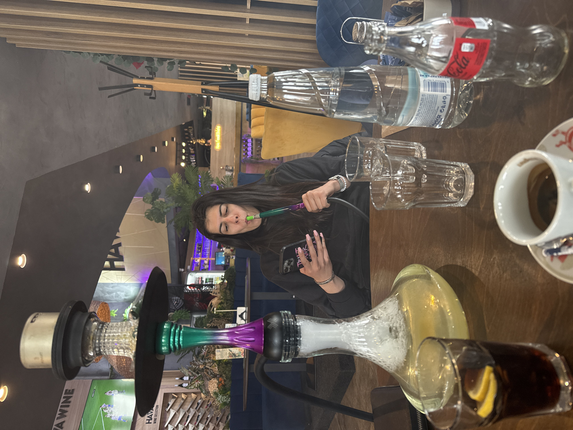 A woman is sitting at a table inside a cafe, smoking a hookah and holding a phone in her hand. She is wearing a black top and has dark hair. On the table, there is a hookah, an empty Coca-Cola bottle, a bottle of water, two glasses, a cup of coffee, and a glass with a dark liquid and lemon slices. The cafe has a modern interior with a mix of wooden and upholstered furniture. In the background, there are plants and a display of wine bottles, as well as other patrons and cafe decorations. A sign visible in the background identifies the establishment as "Hava Wine," a business located in Sofia, Bulgaria.