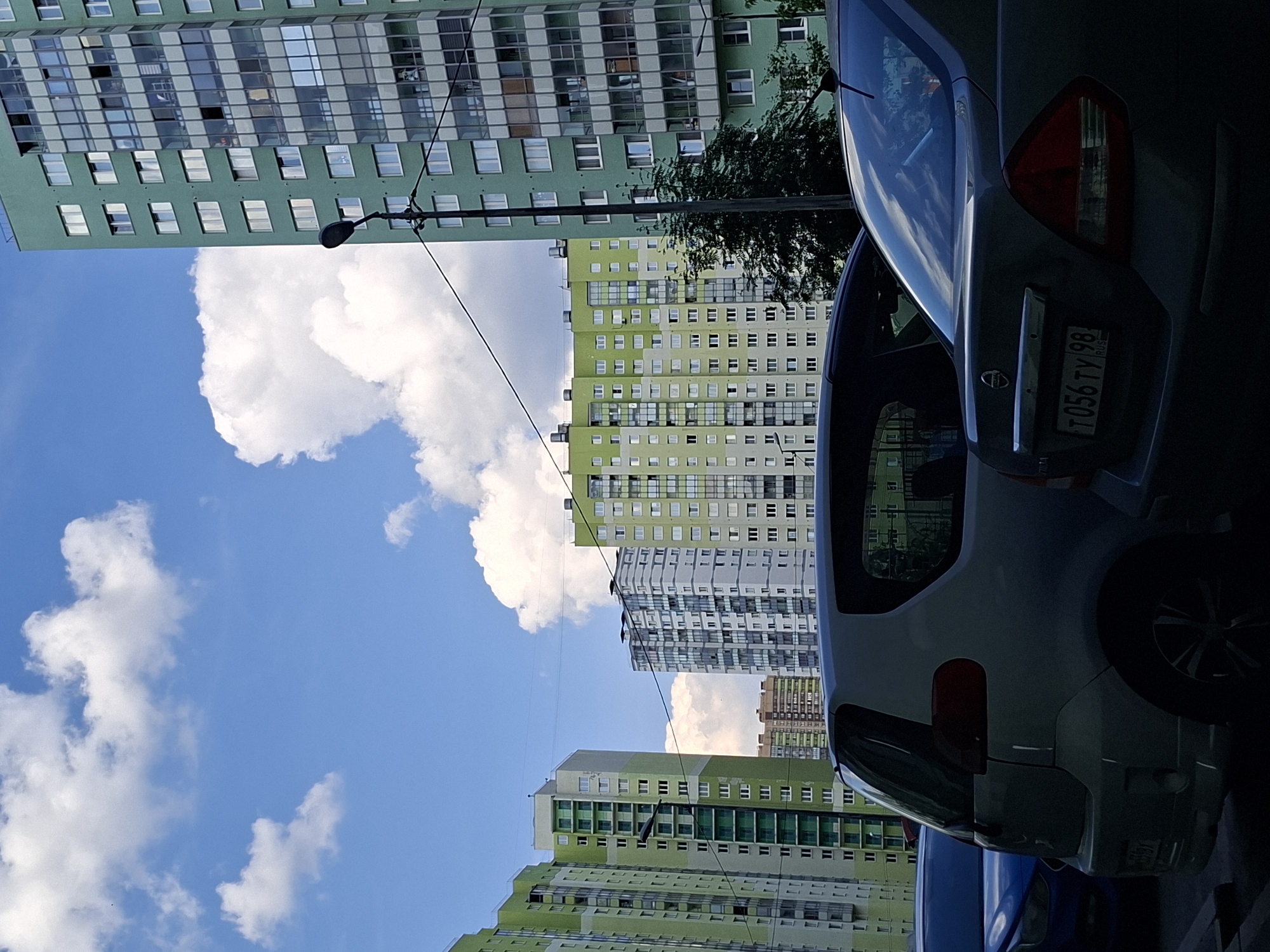 The image captures an urban scene dominated by several tall residential buildings under a bright blue sky with scattered white clouds. The perspective is from ground level, looking upwards, with the buildings extending high into the frame.
In the foreground, on the right side, parts of two parked cars are visible. The closest vehicle is a grey or silver car, seen from its rear and side. Its red taillight is prominent, and the rear window reflects the sky. A Russian license plate is clearly readable, displaying "T056 TY 98 RUS". Behind this car, a section of a blue car is partially visible.
The apartment buildings in the background are multi-story, featuring a modern architectural style. The building on the far left is predominantly green with numerous windows, some reflecting the sky. Other buildings further back and to the right also incorporate green and lighter grey/white panels, arranged in vertical and horizontal sections, indicating a uniform design. These structures appear to be large residential blocks.
A street light fixture on a pole is visible in the mid-ground, positioned against the sky, with thin black wires stretching across the frame. Some green foliage from trees can be seen interspersed among the buildings and cars.
The scene depicts a clear, sunny daytime. The "98" region code on the license plate confirms the location context of Saint Petersburg, Russia. The overall impression is that of a typical residential district in a large city, captured on a pleasant day.