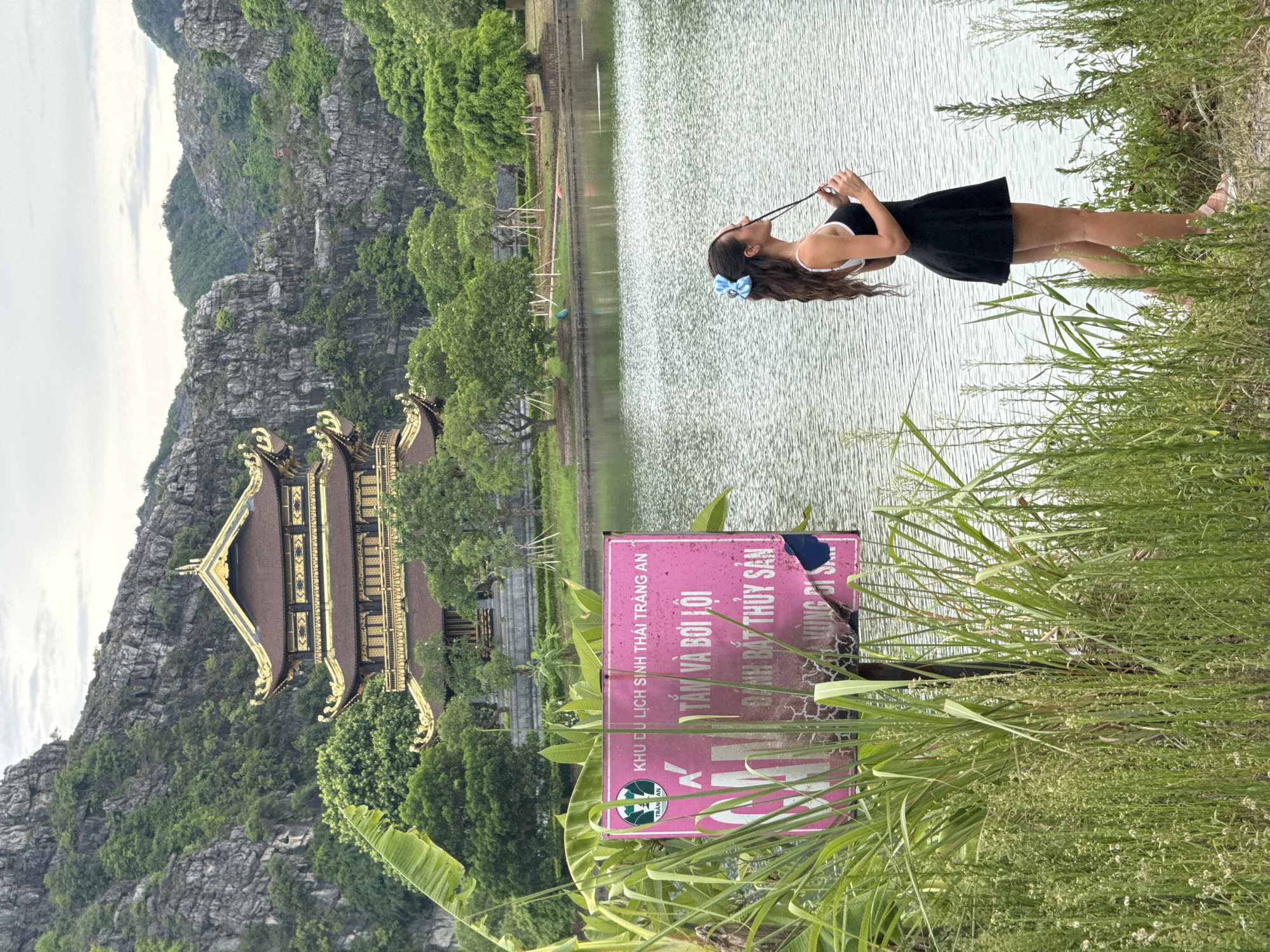 A young woman with long, wavy brown hair and a blue bow stands at the edge of a body of water, holding a thin, dark object. She is wearing a black, sleeveless skater dress with white trim and pale pink sandals. Her profile is visible as she looks towards the water, with her head tilted slightly upwards. The background features a traditional Vietnamese temple with ornate golden roofs, set against a backdrop of lush green trees and dramatic, rocky mountains. In the foreground, a pink sign with Vietnamese text is partially obscured by tall, green grass and reeds. The water is calm with rippling reflections. The sky is overcast with soft clouds.