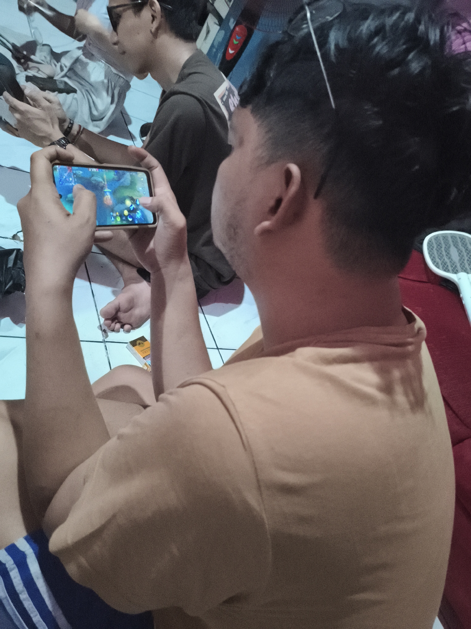 The image depicts a group of young men gathered indoors, possibly in a residential setting in Depok, Indonesia. The primary focus is on two individuals engaged with mobile devices. In the foreground, a person is holding a smartphone horizontally, actively playing a game. The game displayed on the screen features colorful graphics and character icons. Their hands are positioned to interact with the touch screen. To the left of this individual, another person, wearing sunglasses and a dark t-shirt, is also looking at a device, though it's unclear if they are playing a game or engaged in another activity. Their hands are also visible, one resting on their leg and the other partially obscured.
In the background, the environment appears to be a simple room with tiled flooring. Other individuals are present but less clearly visible. One person is seated on the floor, their legs extended. There's a pack of cigarettes on the floor near the person holding the phone. A white electric fly swatter is visible on a red surface in the right portion of the frame. The lighting in the room is somewhat dim, with a blueish cast suggesting artificial light sources. The overall atmosphere suggests a casual gathering of friends.