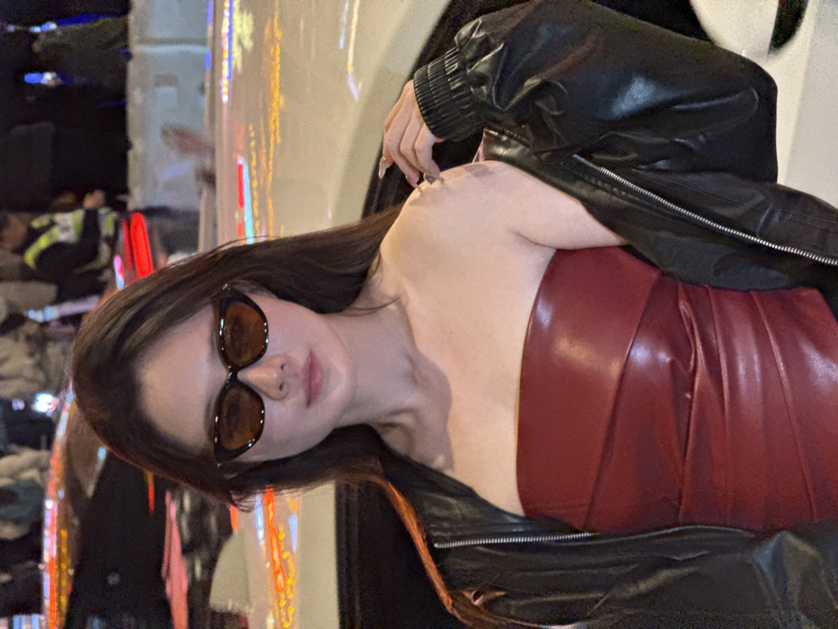 The image depicts a female subject at night in an urban setting identified as Yuzhong District, China. The central figure is an East Asian woman, captured from the waist up, with long, dark hair. She wears dark, cat-eye style sunglasses with amber-tinted lenses. Her attire consists of a strapless, form-fitting, dark red top, which appears to be of a glossy material such as faux leather, featuring a draped or pleated texture across the torso. A black leather jacket is draped over her shoulders; her left arm is partially inserted into its sleeve, while her right hand, exhibiting long, light-colored fingernails, rests on her right shoulder over the jacket. She maintains a direct gaze towards the viewer with a neutral expression. The background is significantly blurred, displaying horizontal streaks of red, orange, and yellow light, indicative of vehicle taillights, brake lights, and other urban illumination sources. Portions of a white vehicle are discernible behind her to the viewer's left, and a dark vehicle to the right. Faint, indistinct figures of other individuals, including one potentially wearing a reflective vest, are visible in the distant background, suggesting a public street or plaza environment.