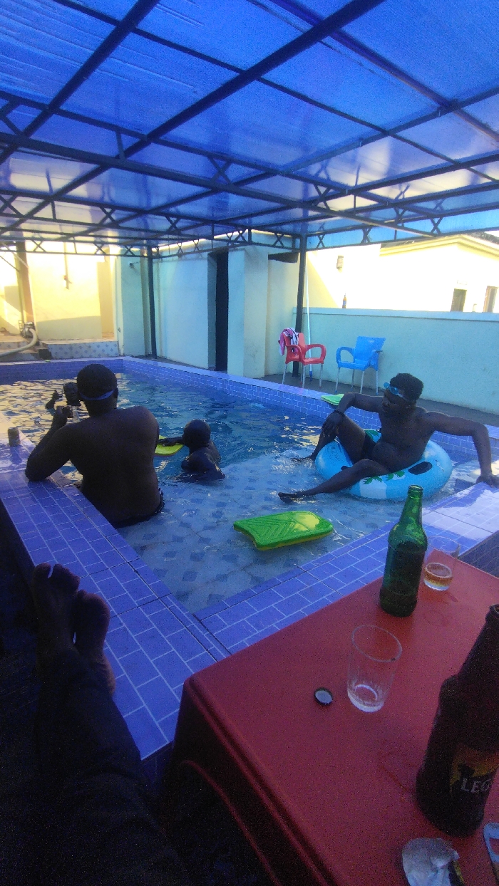 The image depicts an outdoor swimming pool under a blue translucent roof in Ankpa, Nigeria. Three individuals are in the pool; two adults wearing goggles, one sitting in a blue inflatable ring, and a child. A green kickboard floats in the water. A red plastic table is positioned in the foreground, with a green beer bottle, a brown beer bottle, an empty glass, and a glass containing amber liquid on it. One person's feet are visible in the lower left corner of the image. In the background, there are white walls, a window, and two plastic chairs.