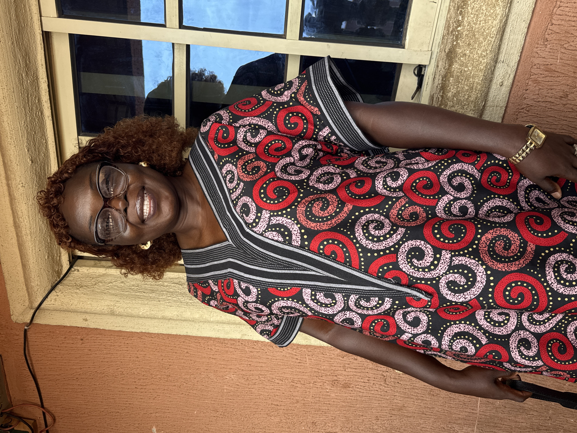 A woman stands in front of a window. She is smiling and wearing glasses. The woman is wearing a dress with red, pink, and yellow patterns, with black and white striped trim. She is also wearing a gold watch. The window has a light-colored frame and reflects the sky. The wall is painted orange and yellow. The location is Onitsha, Nigeria.