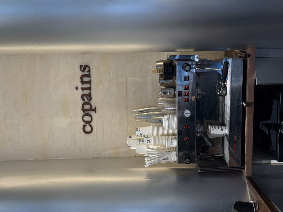This image captures a close-up view of a coffee preparation station within a commercial establishment, specifically the Copains coffee shop in Paris, France.
The scene centers on a professional-grade, two-group espresso machine, crafted from polished stainless steel and black trim, resting on a dark wooden countertop. The machine features several control buttons with red indicator lights, a central pressure gauge, and two group heads. An espresso shot is actively being pulled from the left group head into a white paper cup branded "copains," which has additional French text partially visible. A second portafilter handle, with a black grip, rests on the machine's drip tray next to a crumpled blue cloth.
Above and behind the espresso machine, against a light-colored, textured brick or stone wall, the word "copains" is displayed in raised, dark brown, serif letters. To the left of the sign, a tall, clear container holds numerous white plastic straws. Adjacent to the straws, various stacks of disposable cups are arranged: tall stacks of white paper coffee cups, some branded "copains," alongside a stack of clear plastic cold beverage cups and a stack of white plastic cup lids. To the right, on the machine's surface, several metal steaming pitchers, a black bowl containing a small whisk, and other bar tools are organized. The immediate surroundings consist of reflective, vertical silver-colored panels flanking the counter space, creating a confined, functional work area. No individuals are present in the frame.