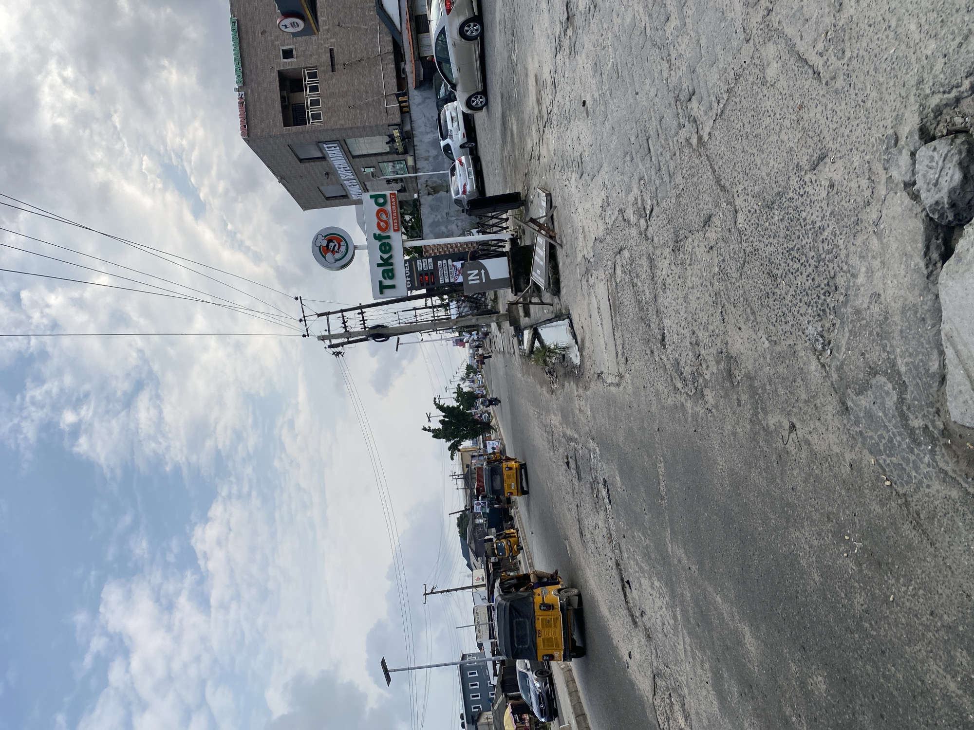 The image depicts a street scene in an urban environment. A paved road with visible cracks and patches runs diagonally from the lower left to the upper right of the frame. Several yellow three-wheeled vehicles, commonly known as tricycles or Keke-Napeps, are present on the road, with two in the foreground and one further in the distance. A silver car is parked on the left side of the road, and other vehicles are visible in the background.
On the right side of the street, a multi-story building with a brick facade stands prominently. A sign for "SPINNEYS SUPERMARKET" is visible on the building. Adjacent to the building, a sign for "Takefood RESTAURANT" is also present, along with a fuel price board and a sign indicating "IN". A utility pole with numerous wires is situated near the Takefood sign. A black metal gate is partially visible in front of the Takefood establishment.
The sky is overcast with a mix of gray and white clouds. Streetlights are visible along the road, indicating it is likely daytime. The overall impression is that of a busy commercial street in a developing city.