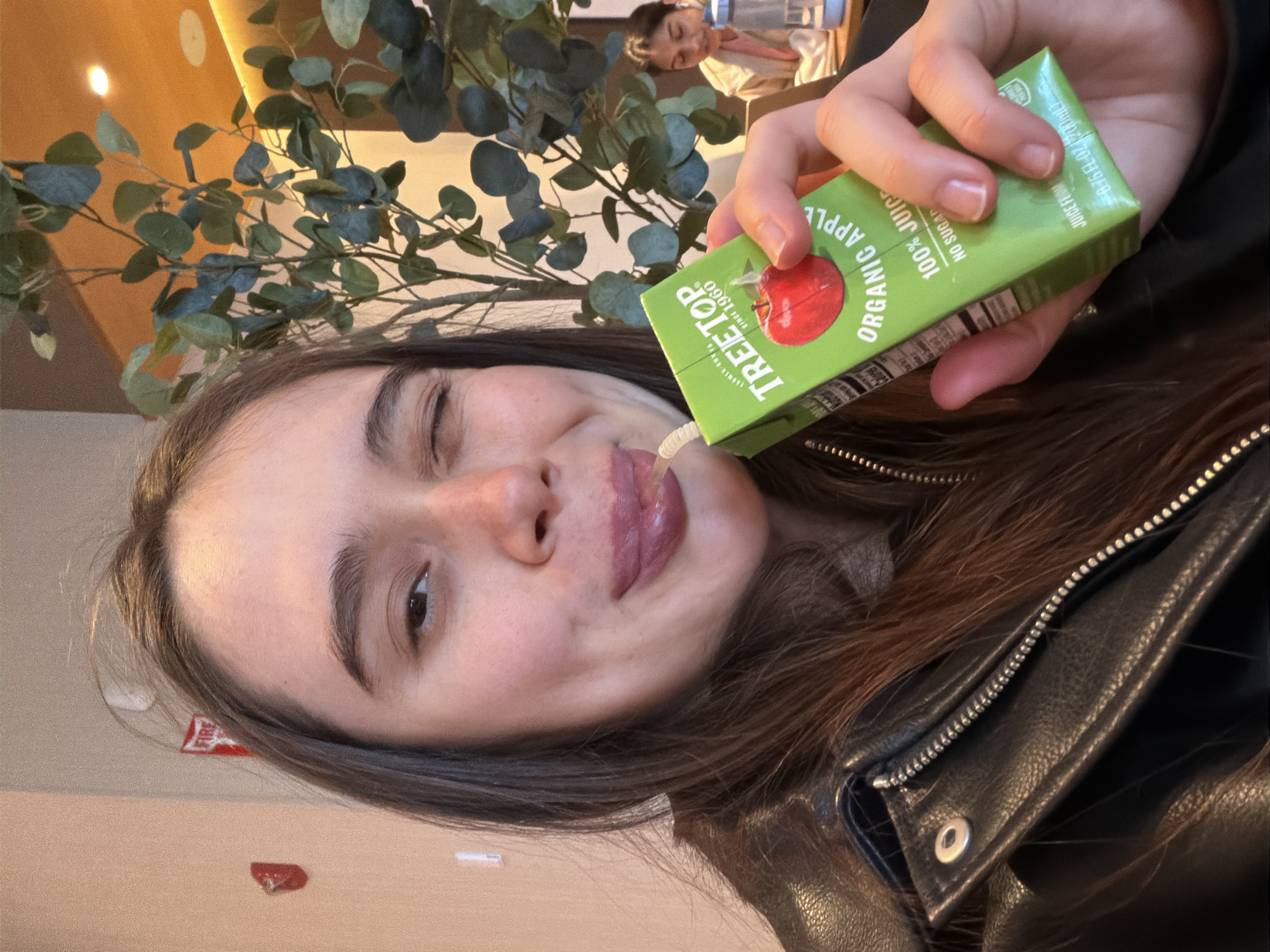 A person, seen from the chest up, is drinking from a green juice box labeled "Tree Top Organic Apple 100% Juice." The person is holding the box up to their lips, with a straw inserted. They are wearing a black leather jacket. A plant with round green leaves is behind the person. In the background, another person is visible. The location appears to be indoors. The city is not directly specified in the image.
