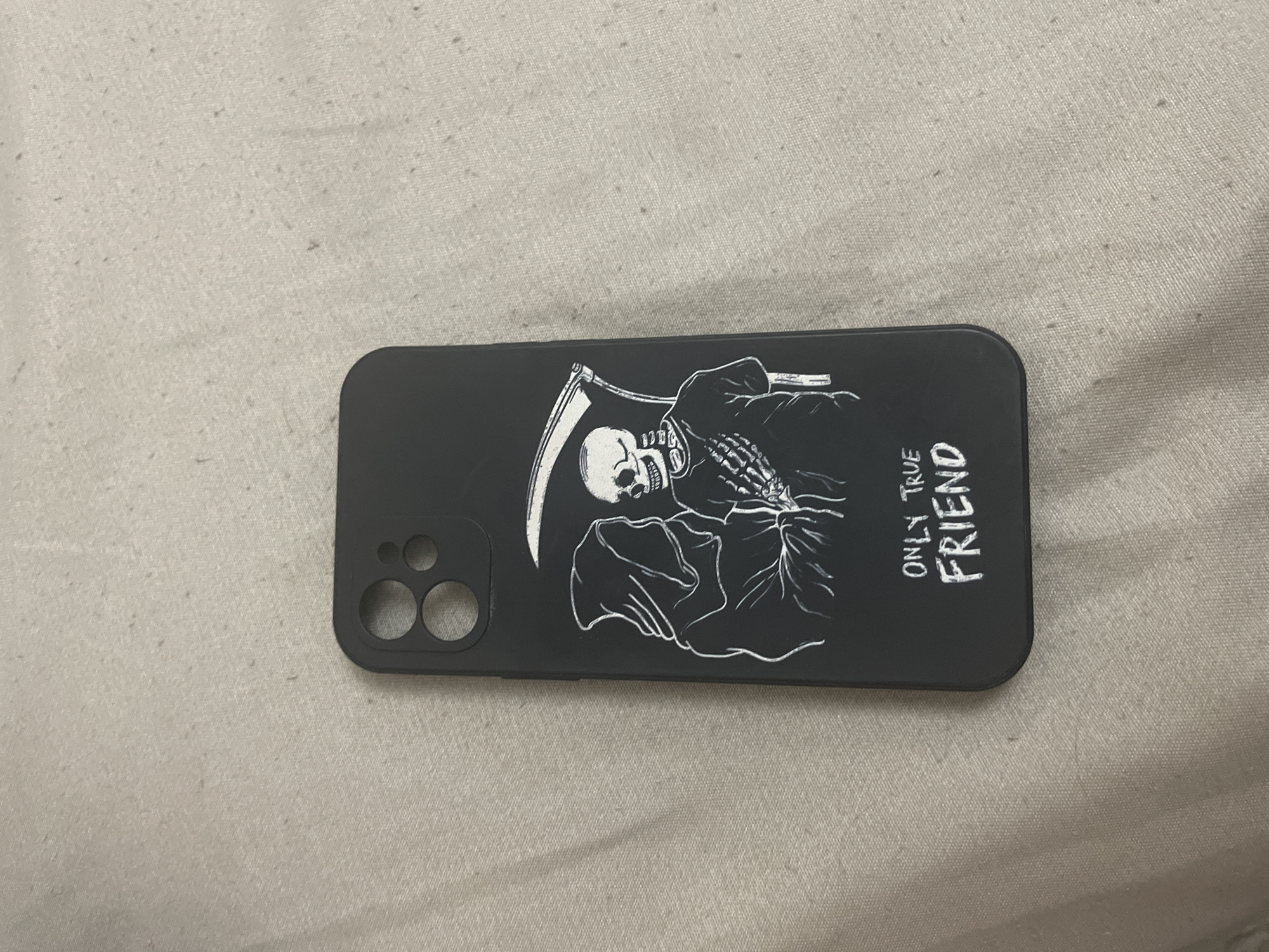 This image presents a matte black smartphone case resting on a light-colored, textured fabric surface within what is specified as a location in Ihtiman, Bulgaria. The phone case features a white line-art illustration depicting two figures: a cloaked, hooded entity resembling the Grim Reaper on the left, and a skeletal figure in a t-shirt on the right, holding a scythe over its right shoulder. The right hand of the cloaked figure and the left hand of the skeletal figure are clasped together at the center. Below the illustration, the phrase "ONLY TRUE FRIEND" is rendered in white, capitalized letters. The upper-left corner of the case incorporates a raised rectangular camera module cutout containing two prominent circular apertures and one smaller auxiliary aperture. The background fabric is a uniform light beige or off-white, displaying a subtle woven texture and scattered small, dark particulates. The scene is evenly illuminated, suggesting an indoor setting.