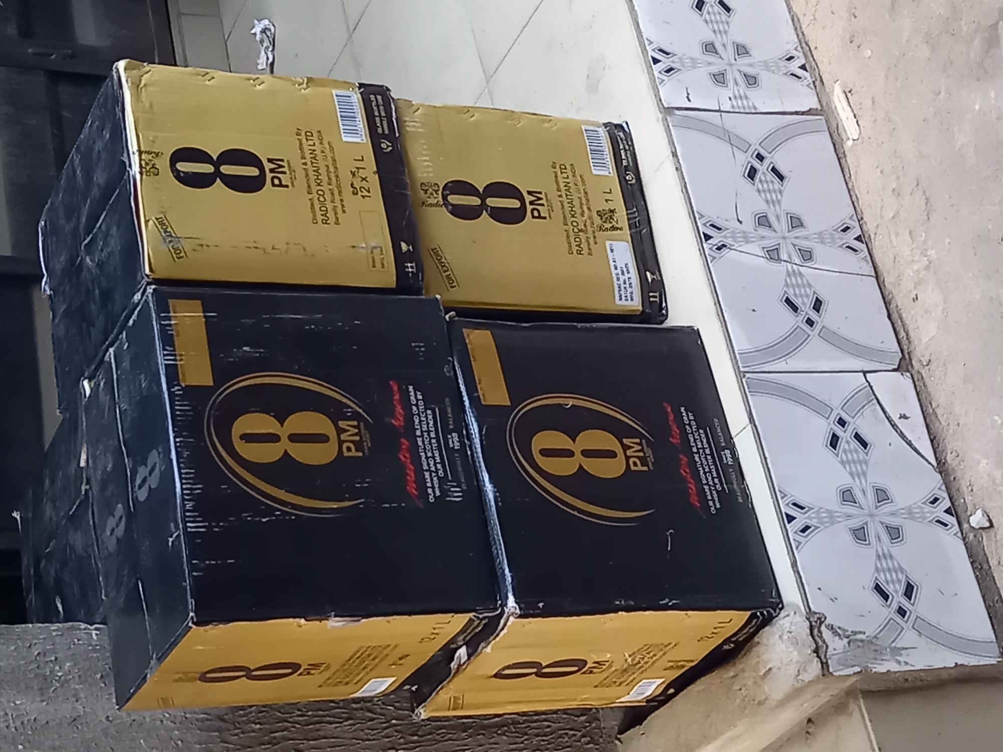 The image displays several cardboard boxes of alcoholic beverages, branded "8 PM," stacked on a floor in Onitsha, Nigeria. The boxes are a mix of golden-yellow and black.
The golden-yellow boxes prominently feature the "8 PM" logo and state they are "Distilled, Blended & Bottled By RADICO KHAITAN LTD" from "Bareilly Road, Rampur (U.P.) INDIA," with the website "www.radicokhaitan.com." These boxes are labeled "12 x 1 L," indicating twelve 1-liter bottles, and some clearly show "FOR EXPORT."
The black boxes also display the "8 PM" logo and contain descriptive text such as "OUR RARE SIGNATURE BLEND OF GRAIN WHISKY AND SCOTCH SELECTED BY OUR MASTER BLENDER" and "1993 BALANCED." One visible label on a black box reads "GLASS BOTTLES HANDLE WITH CARE."
The boxes are arranged in stacks, appearing to be in a storage or transit area. The floor consists of white tiles with a repeating grey and black geometric pattern, bordering a rough concrete or dirt ground. No people are visible, and the scene suggests the storage or handling of these packaged goods.