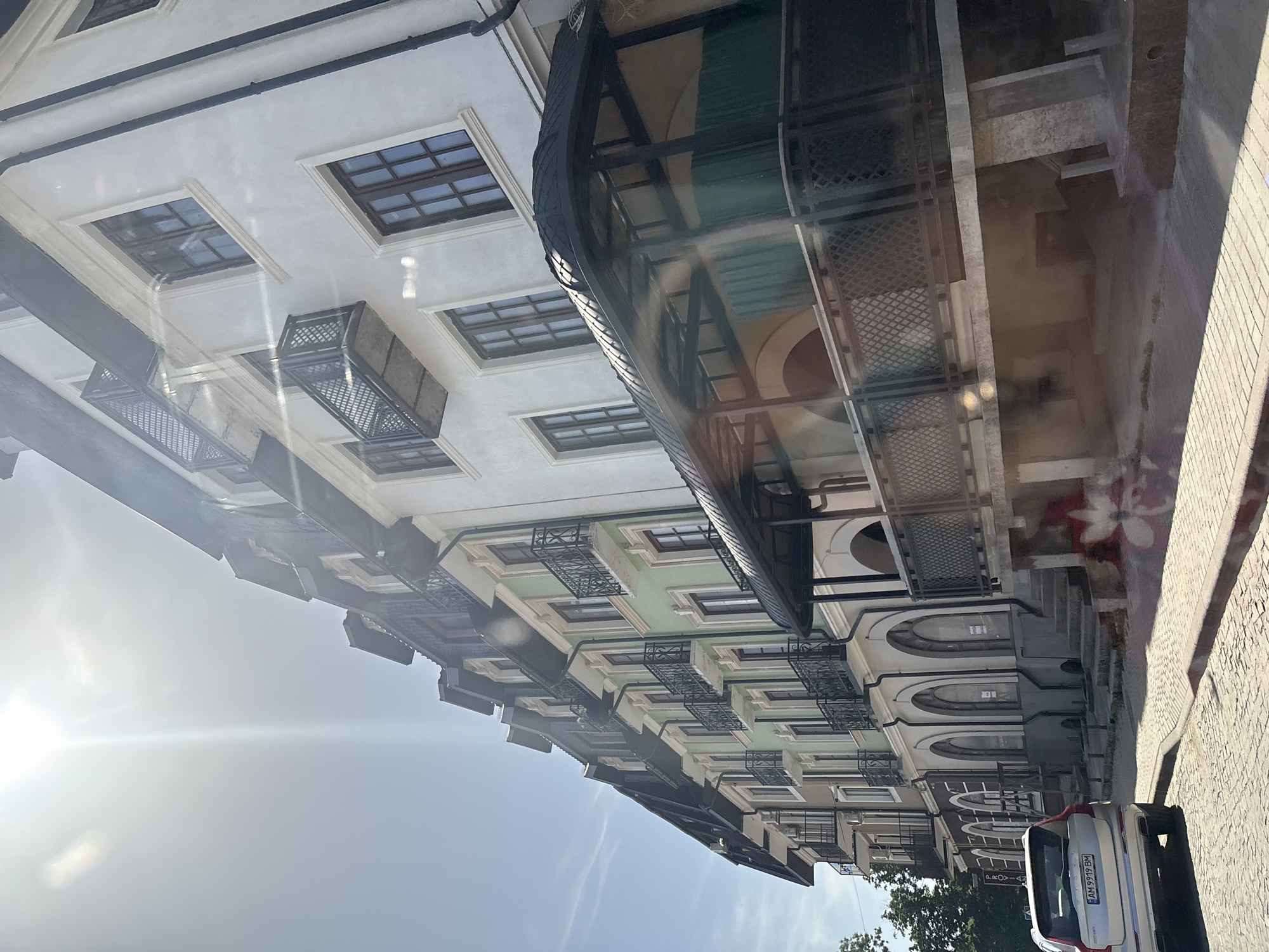 The image shows a multi-story building with a light green facade and a white facade, viewed from a street level. The building features multiple floors with balconies and dark-colored railings and window frames. The sun is shining brightly, casting a lens flare across the upper part of the image. A silver Volvo SUV is parked on the cobblestone street in the foreground. The building's architecture suggests an older European style, possibly in a city center or a historic district.
