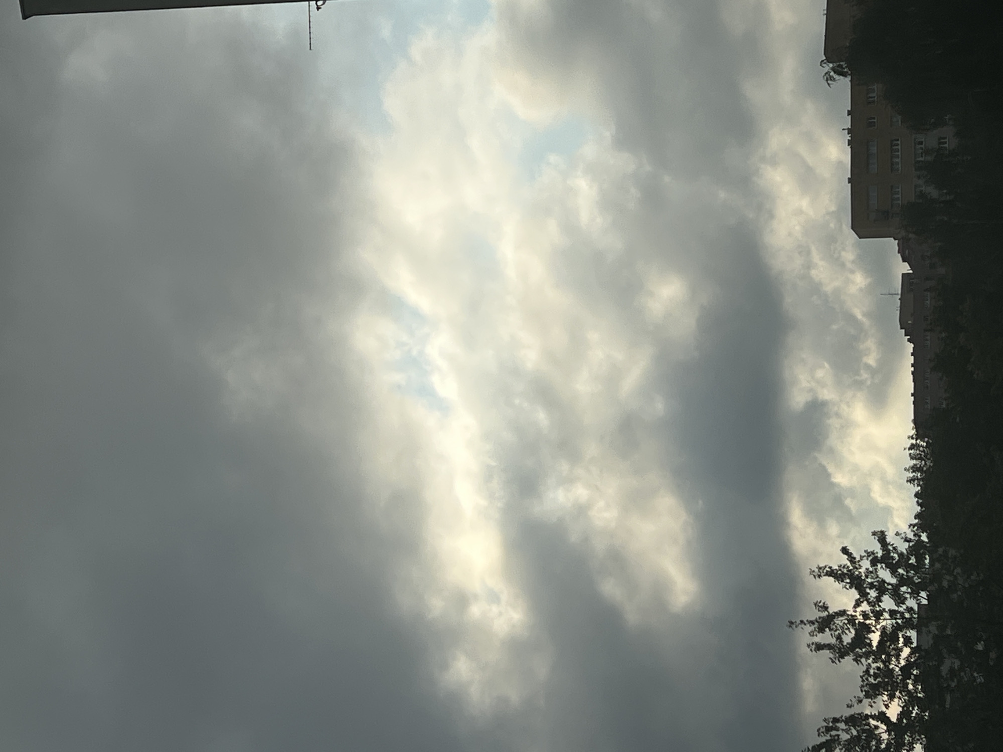 The image depicts a cloudy sky over a residential area in Warsaw, Poland. The clouds are a mix of dark gray and lighter, illuminated patches, suggesting a dramatic sky. In the lower portion of the image, the silhouettes of trees and multi-story apartment buildings are visible against the sky. The architecture of the buildings suggests a typical Eastern European urban landscape. A thin antenna or mast is visible on the far right edge of the image. There are no people or vehicles clearly identifiable in the scene. The overall impression is one of an overcast day in a city.