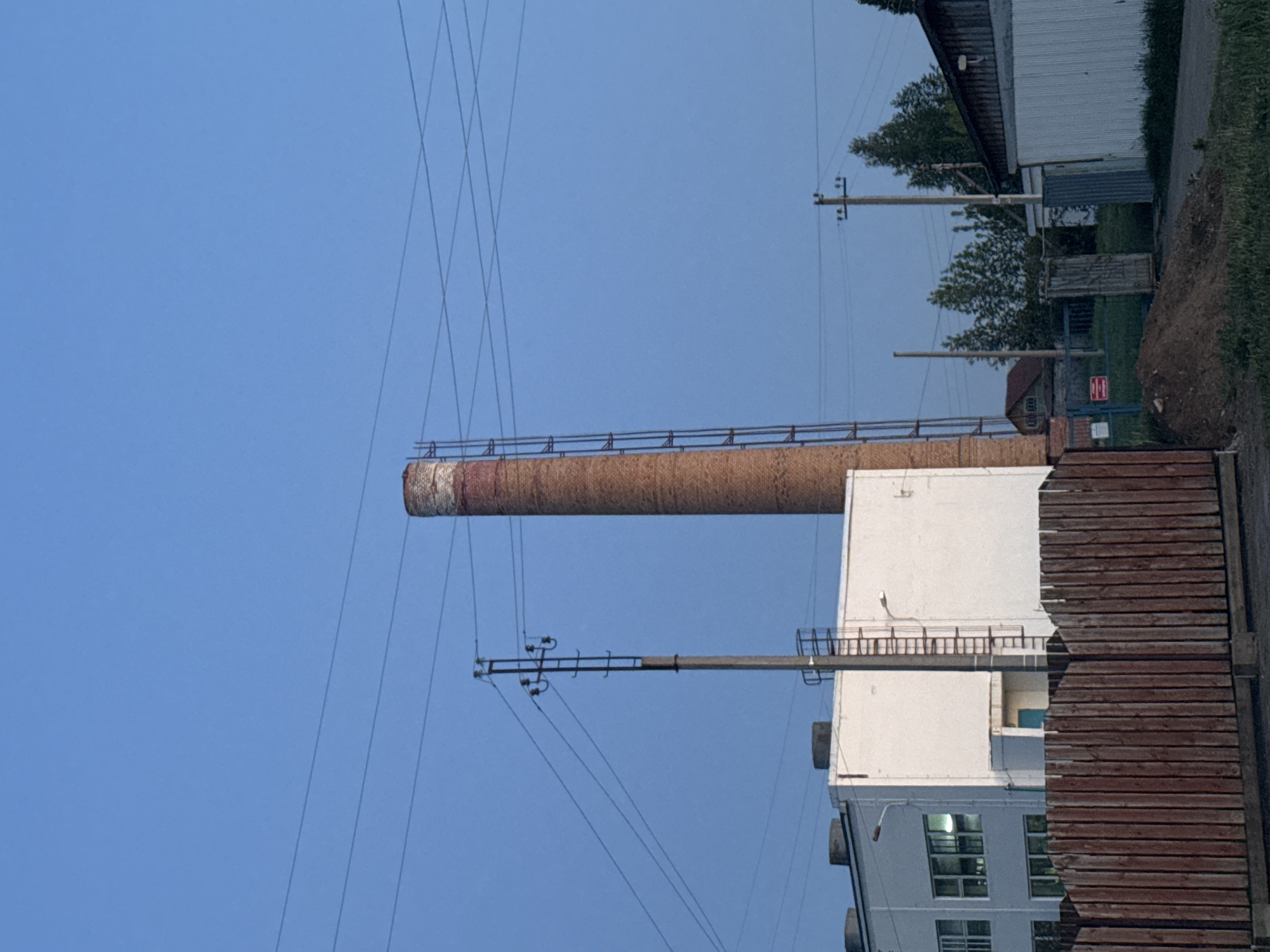 The image shows a brick smokestack connected to a white building in Berezovka, Russia. Power lines run near the smokestack and to a utility pole. The foreground shows a weathered brown wooden fence, and further back, a glimpse of more buildings and vegetation. The sky is clear blue.
