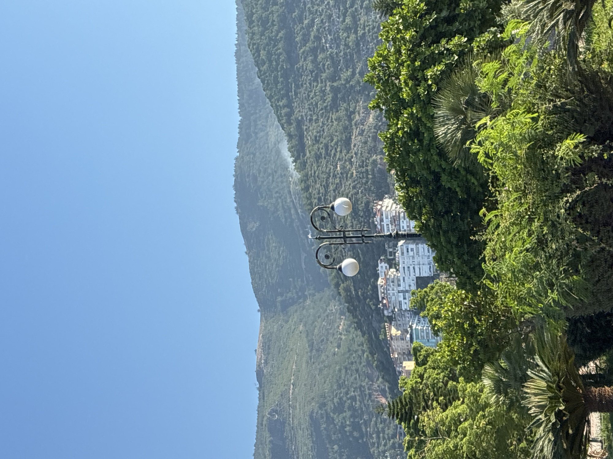 This is a photograph of the city of Alanya, Turkey, on a clear, sunny day. The foreground is filled with lush green trees and palm trees, creating a dense foliage. In the middle ground, several multi-story white buildings with red-tiled roofs are visible, clustered together on a hillside. A street lamp with two white spherical lights stands prominently on the right side of the frame. The background is dominated by a large, green, tree-covered mountain that rises into the clear blue sky. A faint wisp of smoke can be seen rising from the mountainside in the upper right portion of the image.