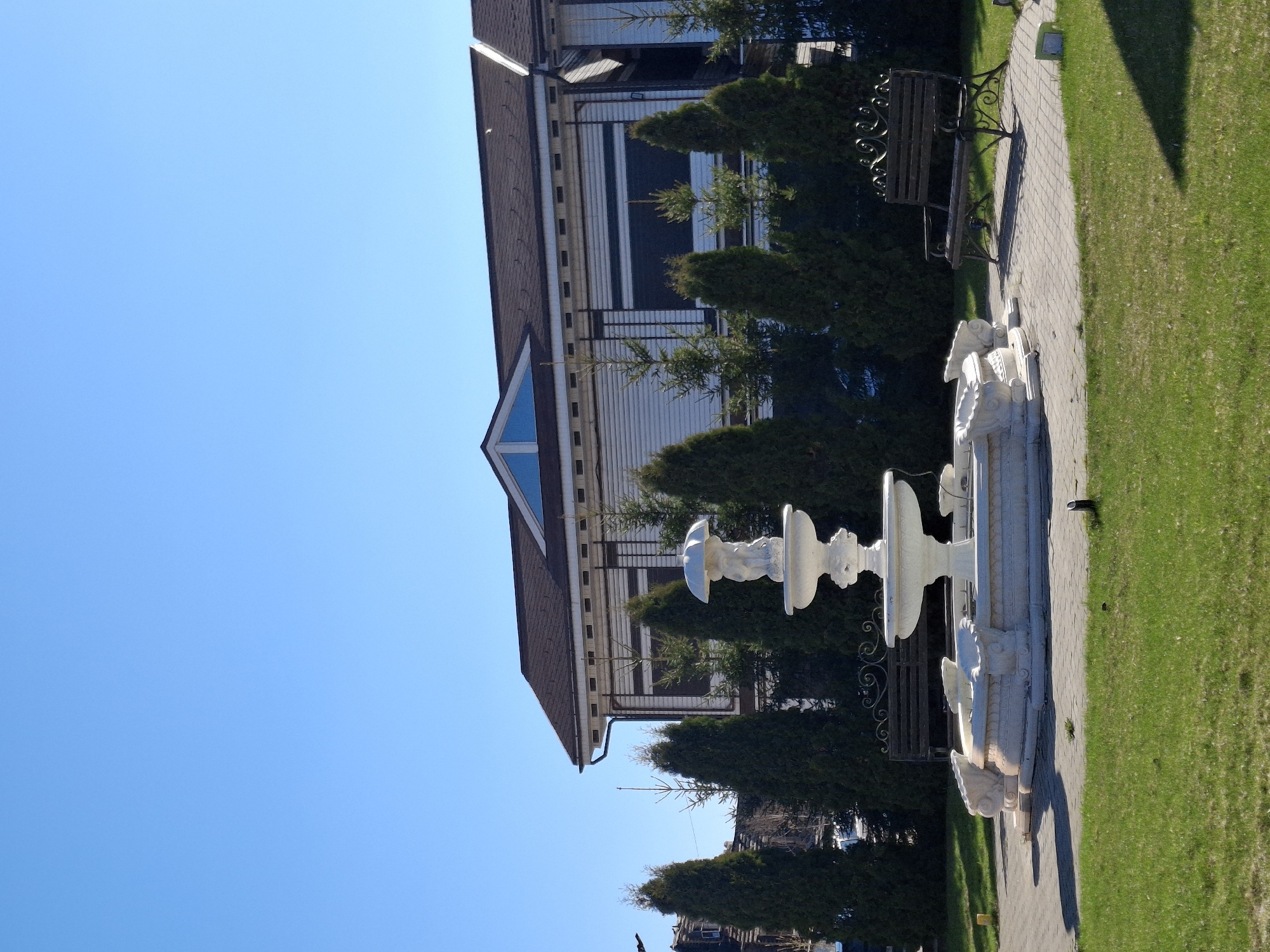 The image shows a two-story building with a brown roof and white siding. A triangular window is visible on the upper level. A white, tiered fountain stands in the foreground, situated on a stone pathway. Adjacent to the pathway is a patch of green grass. A bench sits behind the fountain. A clear blue sky is visible in the background. The image is taken outdoors in what appears to be a park or garden setting. This is likely a site in the city of Sarapul, Russia.