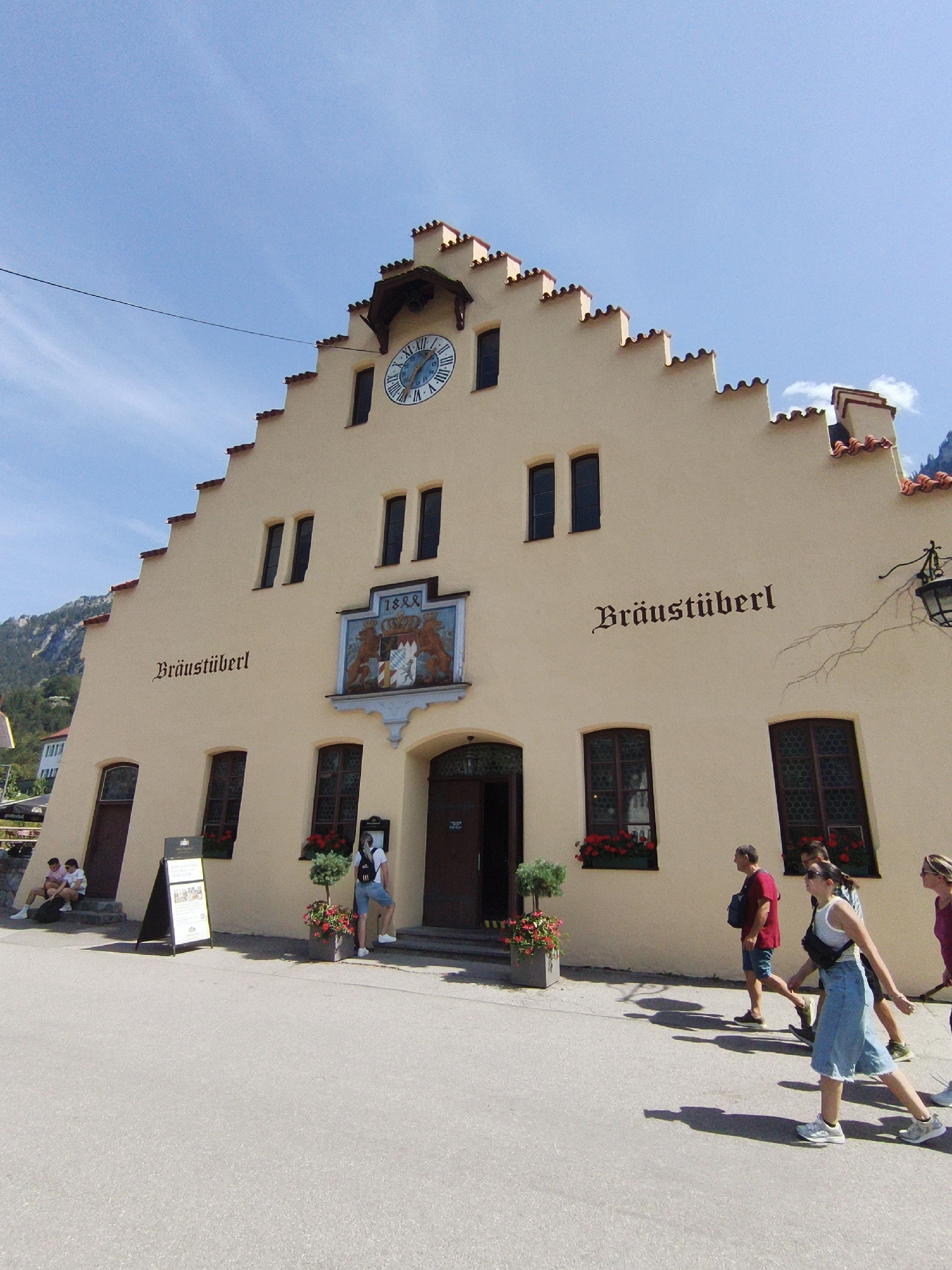 The image depicts the historic "Bräustüberl" building in Schwangau, Germany, on a clear, sunny day. The building has a distinctive stepped gable roof with red tiles and light-colored stucco walls. A large, round clock with Roman numerals is prominently displayed on the facade. Above the entrance, a crest with two lions and a shield is mounted. The text "Bräustüberl" is written in a stylized font on the building's exterior.
Several people are visible in front of the building. In the foreground, four individuals are walking on a paved area. A woman in a blue skirt and white top is in the center, followed by a man in a red shirt and a man in a dark shirt. Further to the left, two individuals are sitting on steps near a standing sign, and another person stands near a potted plant and the entrance. The environment includes a paved walkway, stairs leading to the entrance, and some greenery. The backdrop shows a mountainous landscape partially visible under a blue sky with scattered clouds.