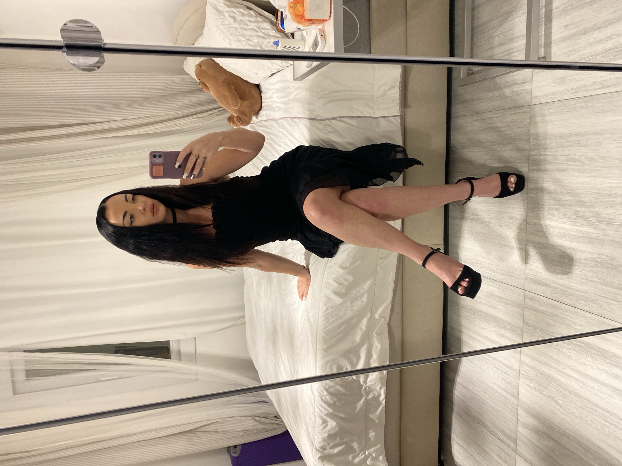 A woman with long dark hair and heavy eye makeup, wearing a black strapless dress and black platform heels, is sitting on a white duvet-covered bed in a bedroom. She is holding a purple smartphone in her right hand and taking a selfie in a full-length mirror. The room has sheer white curtains covering a window on the left and a bedside table with various items on the right, including a plush toy. The floor is tiled with a light gray wood-grain pattern. The overall impression is a posed photograph taken indoors. There are no visible elements that directly indicate the location as Sharm El Sheikh, Egypt.