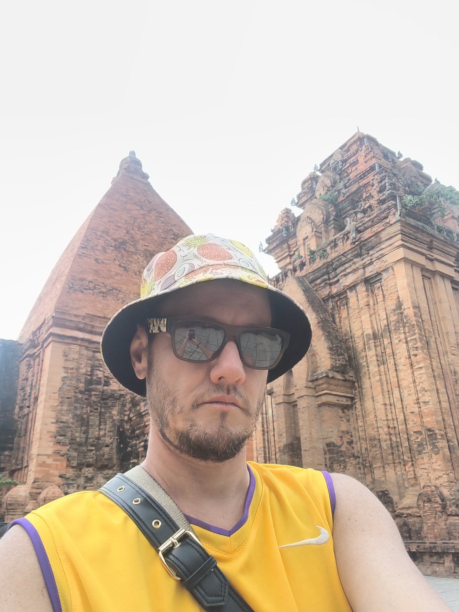 In Nha Trang, Vietnam, a man stands in front of the Po Nagar Cham Towers, a historical complex. He is wearing a yellow tank top with purple trim and a bucket hat adorned with a pineapple pattern. He has on sunglasses and a light beard. A black strap with gold buckles is slung across his shoulder. The Po Nagar Cham Towers, constructed from reddish-brown brick, are visible behind him, with one tower appearing conical and the other more rectangular and ornate. The sky is overcast.