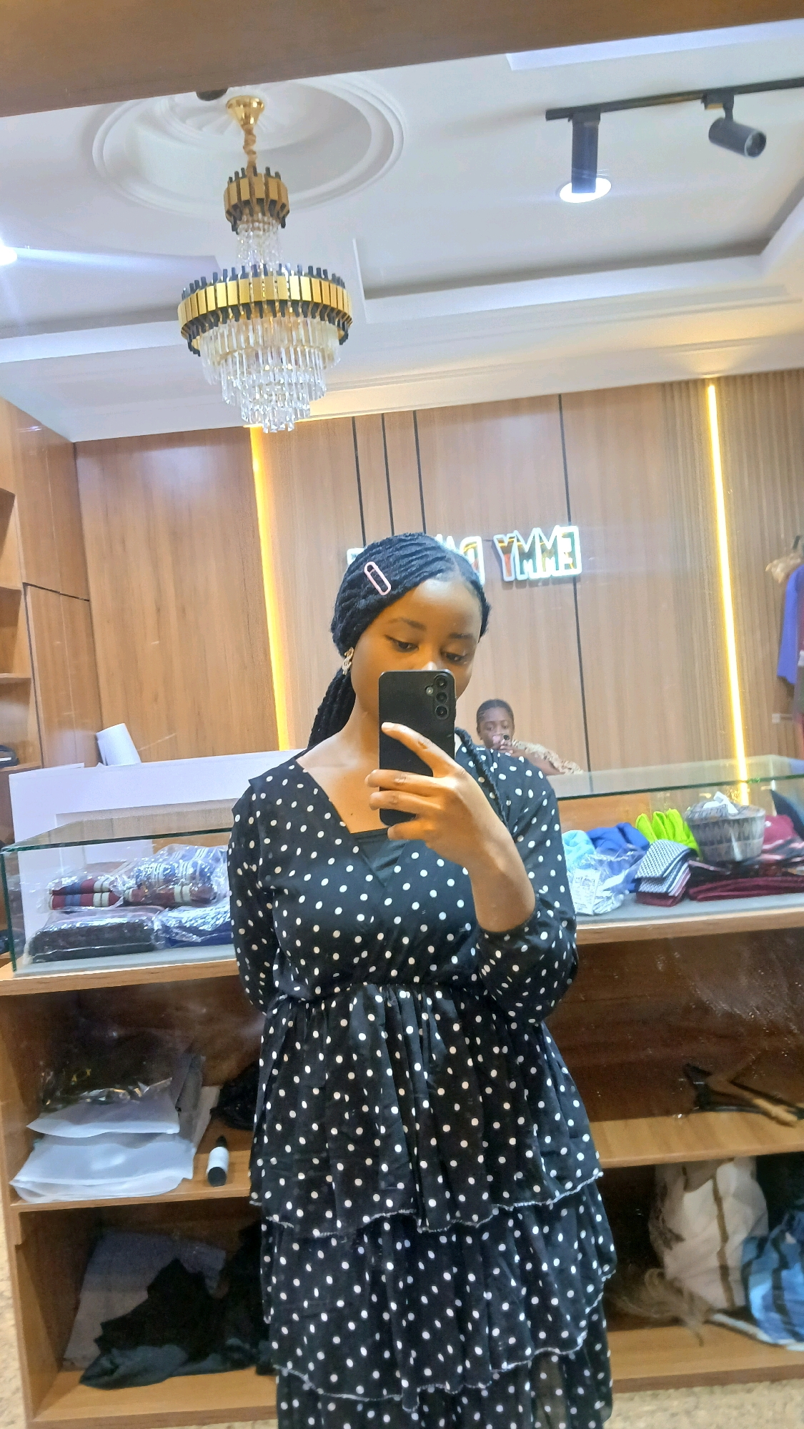 A young Black woman stands in front of a mirror, taking a selfie with a smartphone. She has her hair styled in braids, with a pink clip adorning her left side. She is wearing a black dress with white polka dots, which has a tiered skirt. Behind her, a reflection shows another person, a young Black male, in the background. The environment appears to be a retail store, with shelves filled with clothing and merchandise visible on either side of the woman. A chandelier hangs from the ceiling, and a track lighting system is also present. The background wall features wooden paneling and a neon sign that reads "EMMY DANCE".