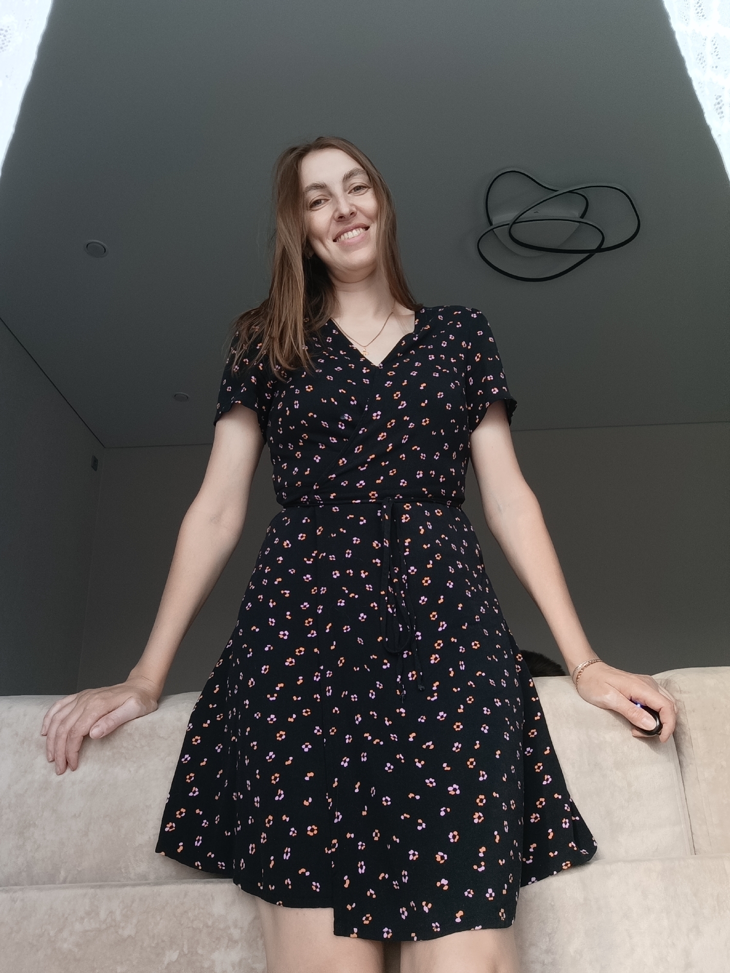 A smiling woman is standing indoors. She has long brown hair and is wearing a black wrap dress with a floral print. She is leaning on a light beige sofa. Above her is a modern ceiling light fixture with intertwined circular elements. The room appears to be in a residential setting, possibly in an apartment, in Chaikovsky, Russia.