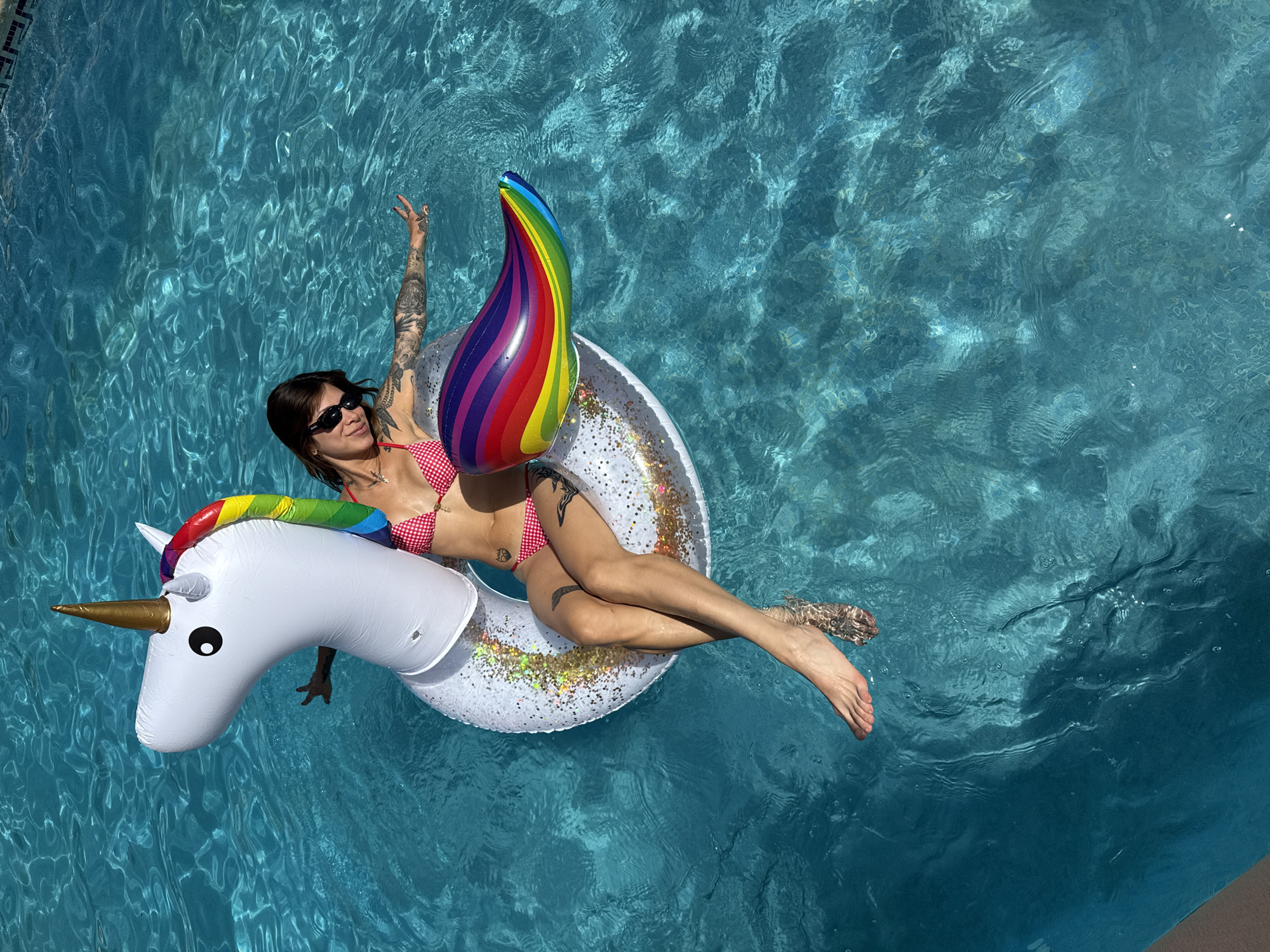 A woman is floating on an inflatable unicorn in a swimming pool in Sant Vicenç de Montalt, Spain. The woman is wearing a red and white checkered bikini and sunglasses. She has tattoos on her arms and legs. The unicorn is white with a rainbow mane and tail. The swimming pool is blue and there are tiles on the bottom. The sun is shining and the water is clear.