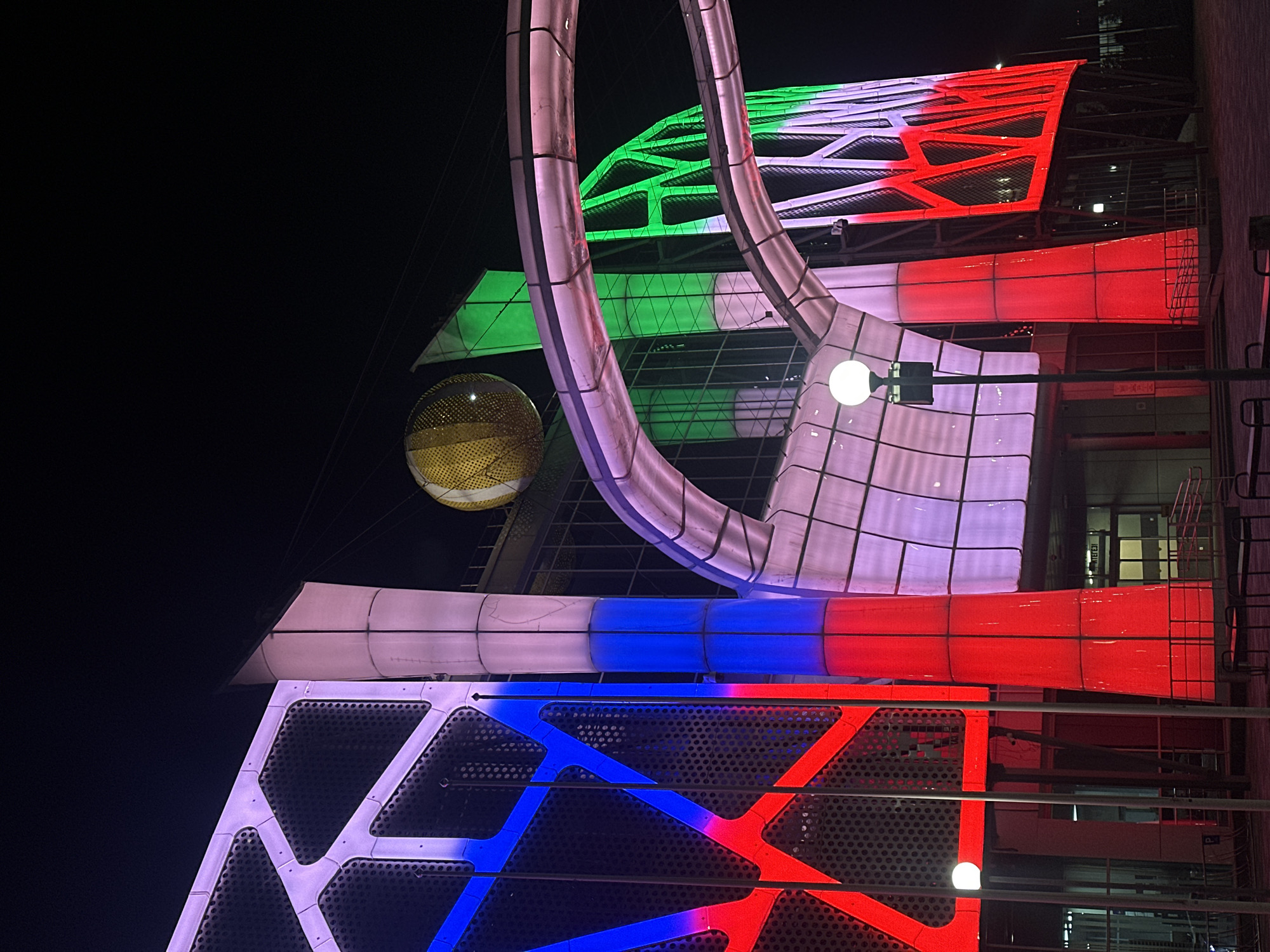 At night, the image shows a colorful architectural installation in Kazan, Russia. The installation has various geometric shapes illuminated in red, white, blue, and green colors. A large circular structure is also present, and a golden, spherical object hangs nearby. The background is dark, indicating that it is nighttime. The building behind the installation has visible windows and architectural elements.