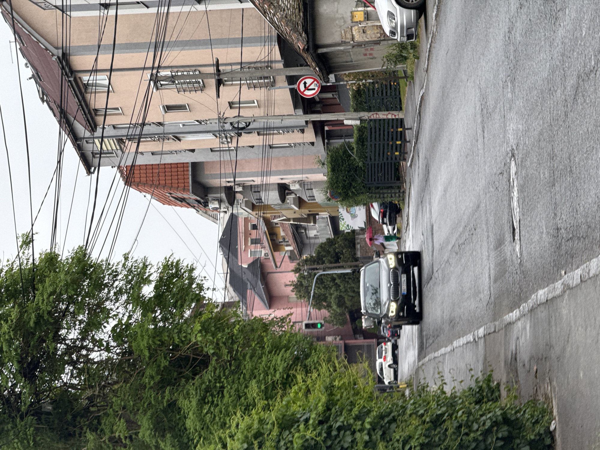 The image shows a street in Vozdovac Urban Municipality, Serbia. The street is wet, suggesting recent rain. Several buildings line the street, of varying colors and heights, along with overhead power lines. A street sign is visible indicating "no U-turn". Several people are visible in the distance, with at least one holding an umbrella. Cars are stopped in the roadway, with a black SUV in the foreground. Trees and bushes line the left side of the street. A traffic light can be seen displaying a green light.