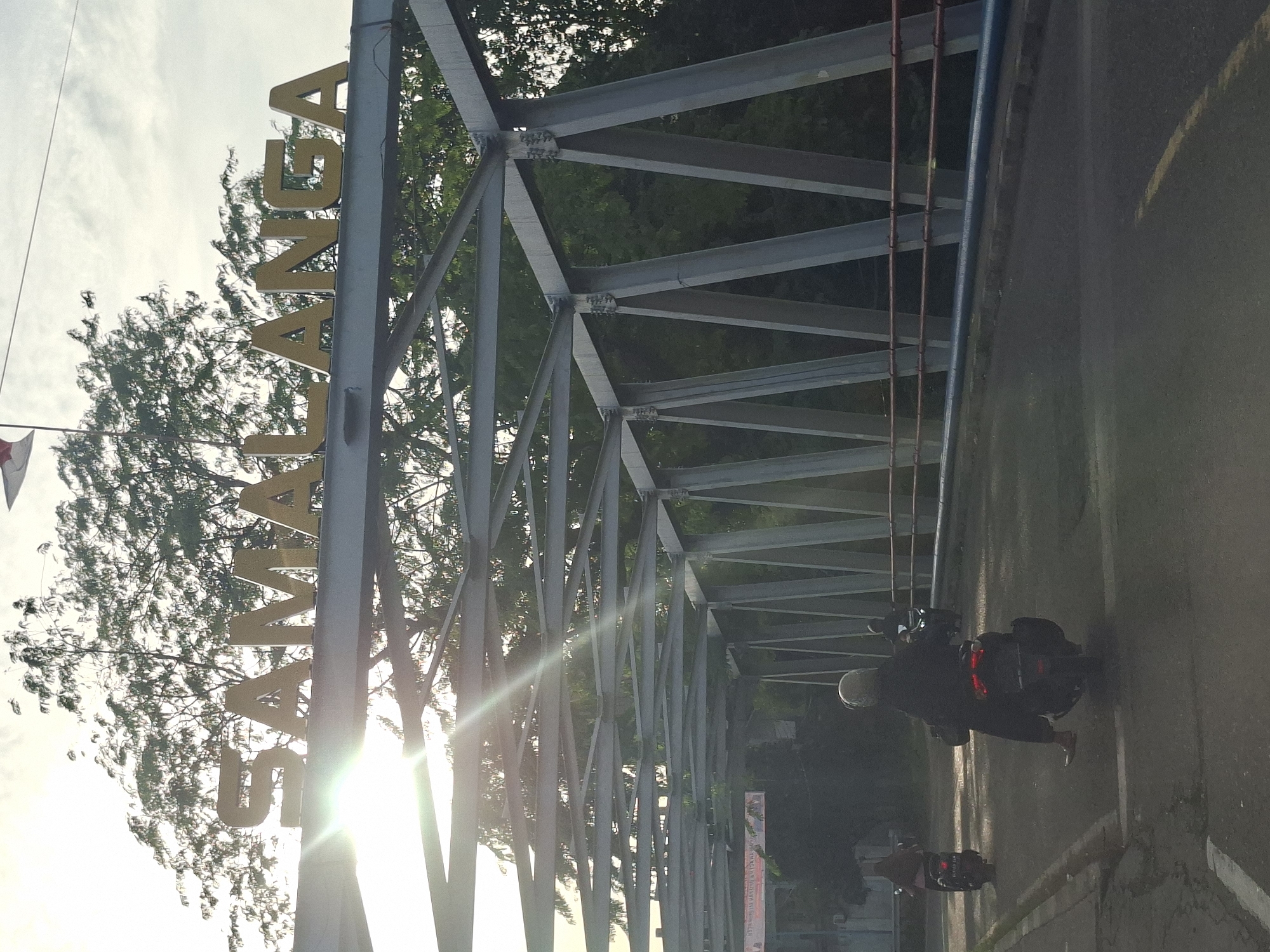 The image shows a bridge over a road in the city of Meurah, Indonesia. The bridge has a large metal sign with the word "SAMALANGA" written in gold letters. The sun is shining brightly, casting rays through the bridge's structure. Several motorcycles are on the road, with riders wearing helmets. In the background, there are trees and a cloudy sky. There is also a flag visible on a pole in the upper right corner. The overall scene appears to be a typical day in the city.