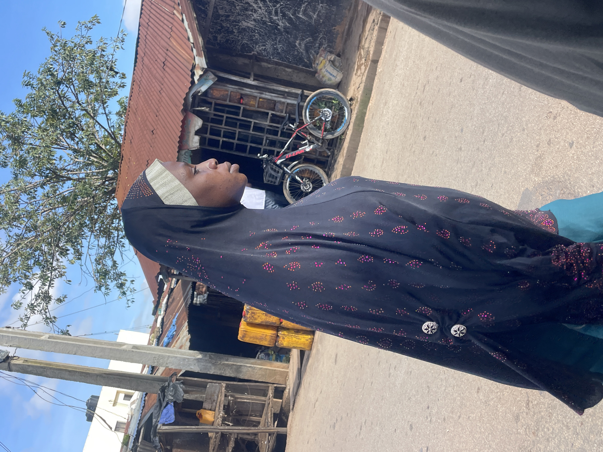A young woman wearing a dark hijab with a light-colored band and pink and gold embellishments walks down a paved street in Jos, Nigeria. She is captured in profile, facing to the right. The hijab features two decorative silver buttons on the left side. She is wearing a teal garment underneath the hijab. In the background, there are several dilapidated structures with corrugated metal roofs and wooden stalls. A bicycle with a black frame, red handlebars, and blue rims is leaning against one of the stalls. Yellow barrels are stacked near a stall, and a white building with a water tank on its roof is visible further back. Utility poles and wires are present in the scene. The sky is blue with scattered clouds. The overall impression is that of a street scene in a developing urban area.