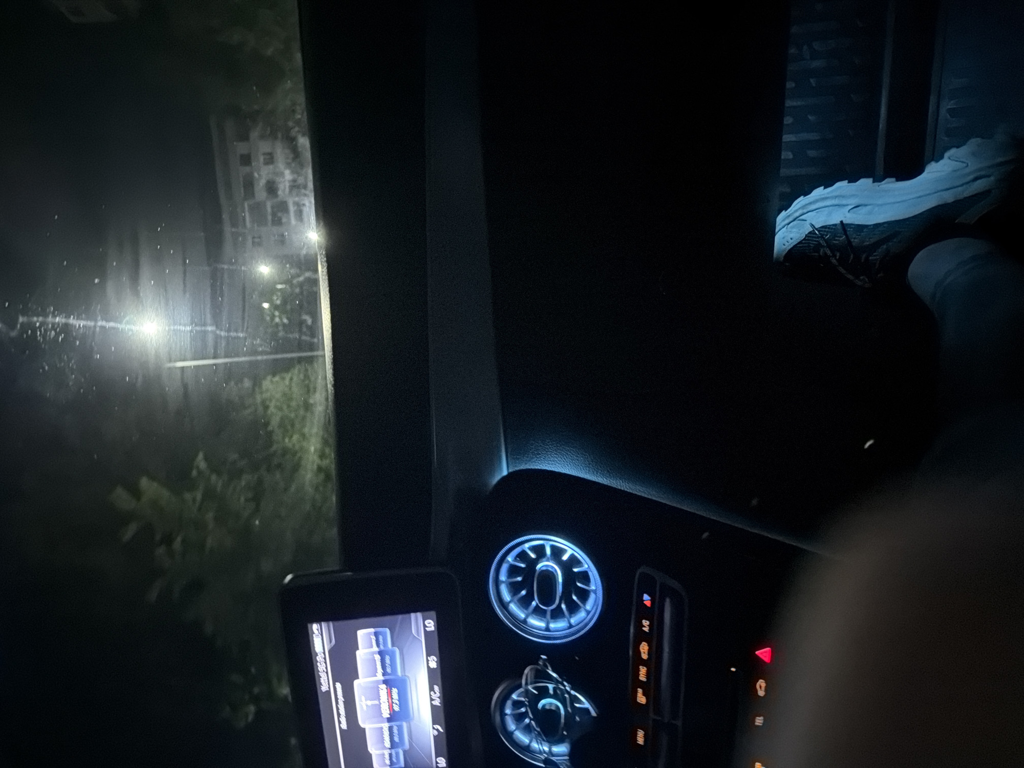 The image captures an indoor-outdoor scene at night, taken from the passenger seat of a vehicle. The car's interior is visible on the right, while the view through a window, possibly the front windshield or passenger side window, is on the left. The outdoor scene is oriented vertically within the frame, as if the photo was taken with the camera rotated.
Inside the vehicle, a person's leg and foot, clad in a light-colored sneaker with dark accents, rest on what appears to be the dashboard or passenger footwell. The car's dashboard features several illuminated elements: two prominent circular air vents with a cool blue backlighting, and a central console with various buttons. A red triangular button, likely for hazard lights, is clearly visible, along with other illuminated buttons labeled "A/C", "SYNC", "MENU", and "TEL". To the left of the central console is a display screen, possibly an aftermarket unit, which shows text including "Virtual SC-PC" and "Radio Station." Further down, radio station frequencies like "97.5MHz" and "98.7MHz" are visible, with "VERONIKA" highlighted, alongside climate control indicators such as "AC/C", "LO", and "S5".
Outside the car, the scene is dark and appears to be raining or heavily misty/foggy, with streaks of light on the wet window surface. Vertically aligned streetlights illuminate parts of the environment. A multi-story building, possibly residential or commercial, is dimly visible in the distance, also vertically oriented in the frame. Some dark foliage or trees are present closer to the road. The overall atmosphere is dark and somewhat blurred by the weather conditions. The location context is Shumen, Bulgaria.