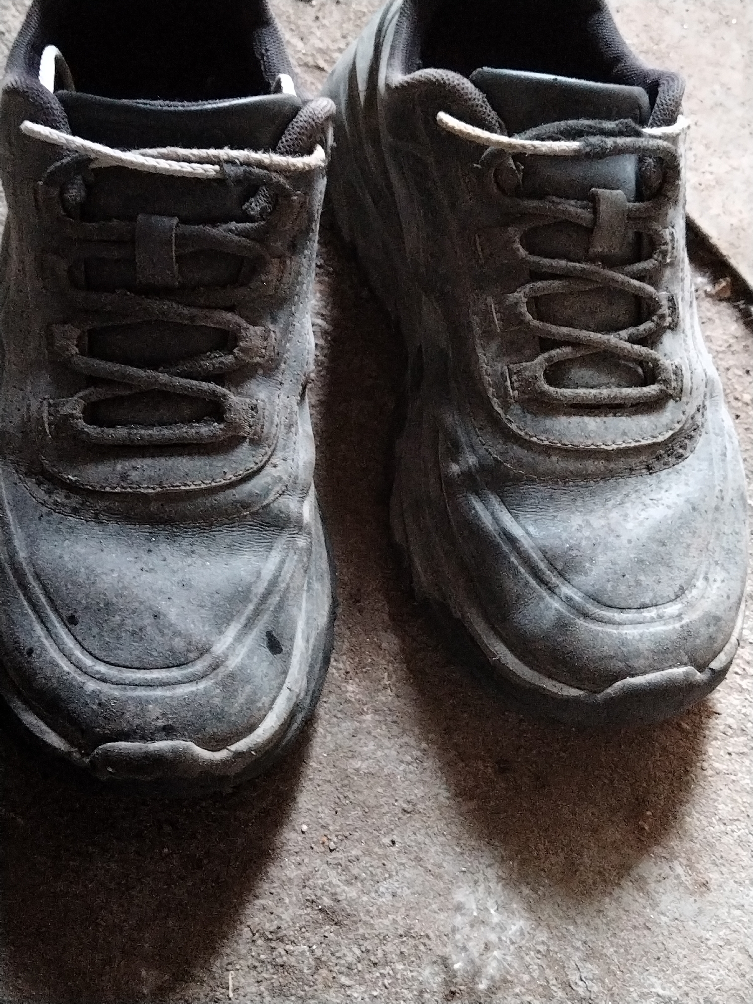A close-up, overhead shot shows a pair of heavily worn, dark gray work boots sitting on a rough, concrete surface. The boots are laced with light-colored laces and appear to be covered in a fine layer of dust or dirt, suggesting recent use in an outdoor or industrial environment. The left boot is slightly closer to the camera and more in focus than the right boot. Shadows are cast to the right of the boots, indicating a light source from the upper left. The background is the textured concrete, with some fine debris visible. No people are present in the image. The scene is devoid of any specific Pargolovo, Russia landmarks or indicators.