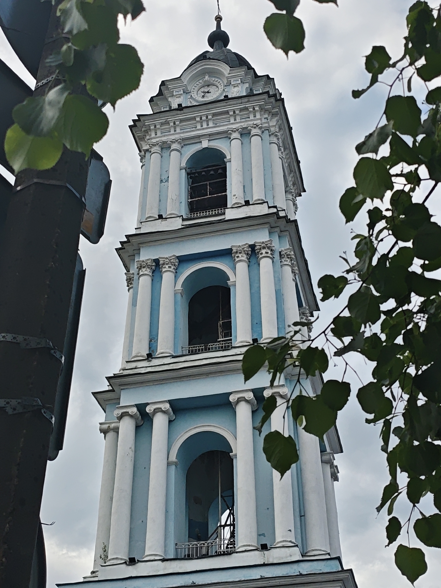 The image is a low-angle shot of a blue and white three-tiered bell tower with white columns and arches. The tower is topped with a small black dome and cross. There is a clock on the top tier of the tower. The tower is located in Noginsk, Russia. In the foreground, there are green leaves from a tree branch on the right and a dark green pole on the left. The sky is overcast.