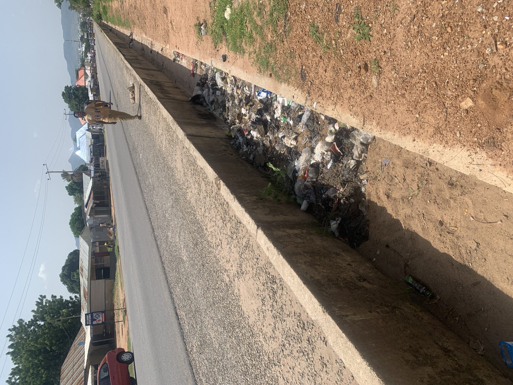 A man walks along a paved road in Jalingo, Nigeria. The road is bordered on one side by a concrete drainage ditch filled with garbage, and on the other side by a dirt path and a line of buildings. The buildings appear to be shops or residences, with corrugated metal roofs. Trees are visible in the background, along with hills in the distance. The sky is clear and the sun is shining. The man is wearing a light-colored t-shirt and pants. He is walking away from the camera. The drainage ditch contains a variety of trash, including plastic bags, bottles, and food wrappers. A red car is parked on the side of the road near the buildings.