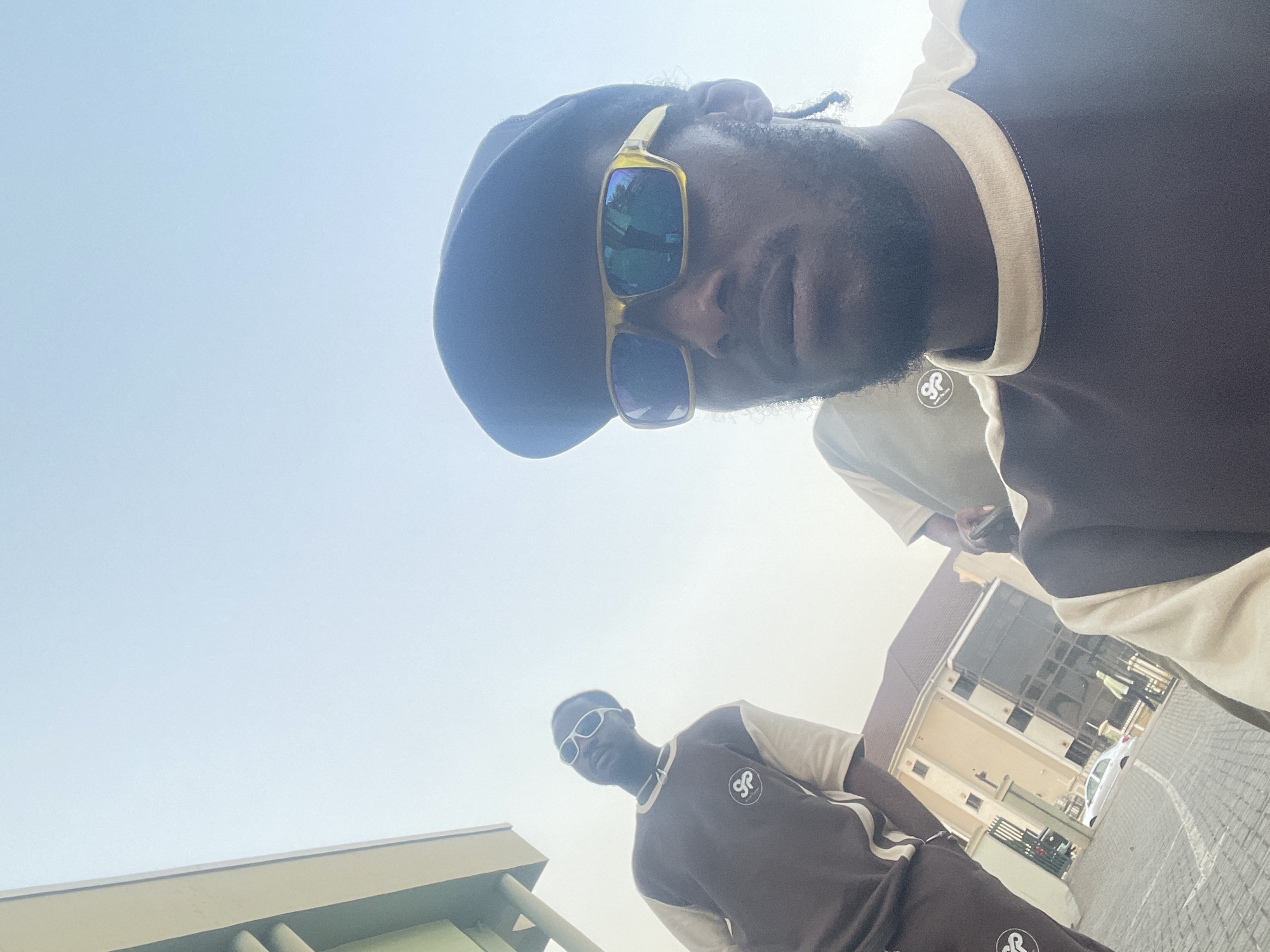 The image, captured from a low-angle perspective, features two men posing outdoors against a bright, clear daytime sky in what appears to be an urban or suburban setting in Abuja, Nigeria.
The dominant subject in the foreground, positioned at the top right, is a man with dark skin and a beard, looking directly towards the camera. He wears a dark cap or beret and distinctive gold-framed sunglasses with reflective lenses. His attire consists of a dark brown shirt or sweatshirt with light beige trim around the collar and sleeves. A circular logo, featuring intertwining letters that resemble "CR" or "GR," is clearly visible on his left chest.
Further in the background, towards the bottom left, stands a second man. He also has dark skin and wears sunglasses with white frames, along with an outfit identical in style and color to the foreground man's—a dark brown top with light beige accents and the same circular logo on his chest. Both men appear to be dressed in matching athletic or casual wear.
The setting includes architectural elements: a light-colored building with pillars is partially visible on the far left, and a modern, multi-story building with dark, reflective windows and a dark roof stands in the background on the right. A white car and paved ground with white markings, suggestive of a parking lot or street, are also visible. The bright light and clear sky indicate it is mid-day, with strong sunlight creating reflections on the sunglasses. The image captures a posed moment, highlighting the two individuals and their coordinated attire.