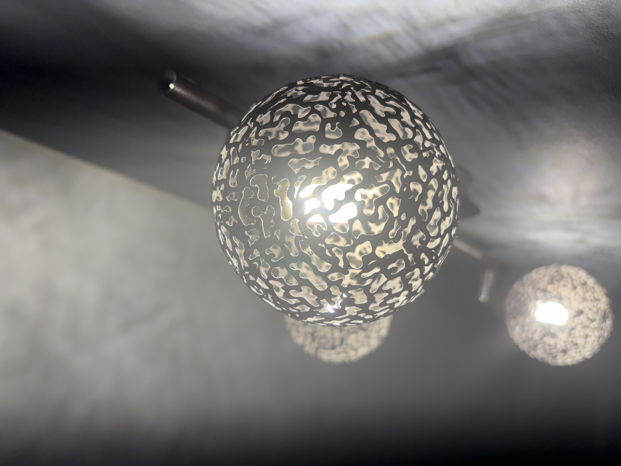 The scene depicts a close-up, upward-angled view of a multi-sphere lighting fixture located within an interior space in Chaikovsky, Russia. The primary subject is a large, illuminated spherical light fixture, prominently positioned in the mid-right of the frame. This sphere features a dark metallic exterior with numerous irregularly shaped, organic perforations, through which a bright white light source is visible. Light reflects brightly off the inner surfaces of the sphere and casts distinct, patterned shadows onto a textured, light-grey wall surface to the right. Behind and below the primary sphere, at least two additional, smaller, spherical luminaires of similar design are visible, progressively out of focus and also illuminated. These spheres are mounted on a dark, slender, horizontal rod or arm that extends diagonally from the upper right towards the lower left of the frame. The background consists of a textured, light-grey surface on the right and upper portions, fading into a dark, indistinct area on the left. No individuals or specific actions are depicted.