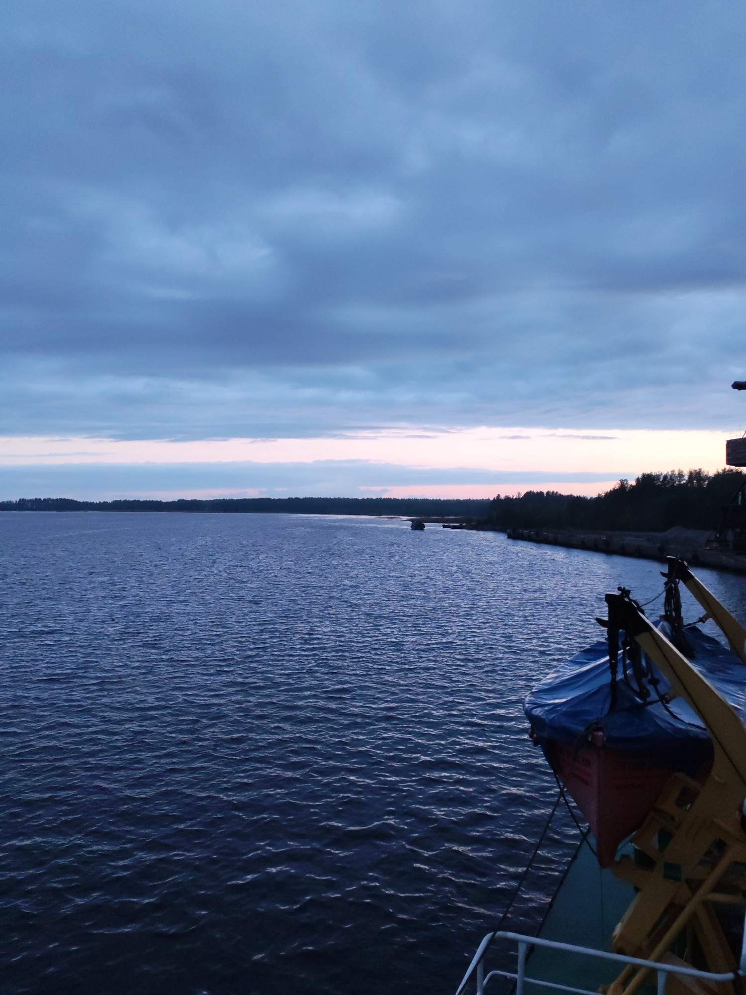 The image depicts a view from a vessel on a body of water, possibly a river or a lake, under an overcast sky with streaks of pink on the horizon suggesting twilight. The location is identified as Shalsky, Russia. The water is dark and choppy, with visible ripples across the surface. A wooded shoreline is visible in the distance. A partially obscured orange lifeboat covered in blue is affixed to the side of the vessel. The deck railing of the vessel is visible in the foreground.
