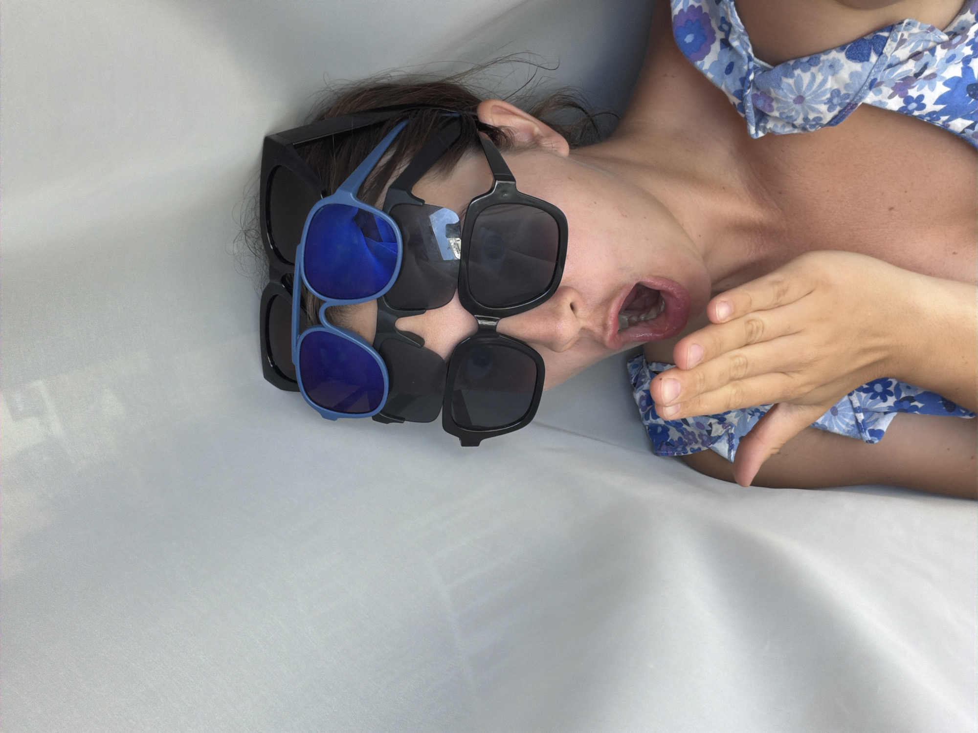 This is a vertical, close-up, eye-level shot of a person lying on a white, textured surface. The person's face is partially obscured by multiple pairs of sunglasses stacked on their head, and one hand is raised to their face. They appear to be wearing a blue floral print top. Their mouth is open, and their eyes are not visible due to the sunglasses. The lighting is bright and diffused, suggesting an indoor setting. The background is a soft white, likely a sheet or fabric. The overall impression is playful or whimsical. There is no visible text in the image. The location context of Sochi, Russia, does not provide any specific visual cues within the image itself.