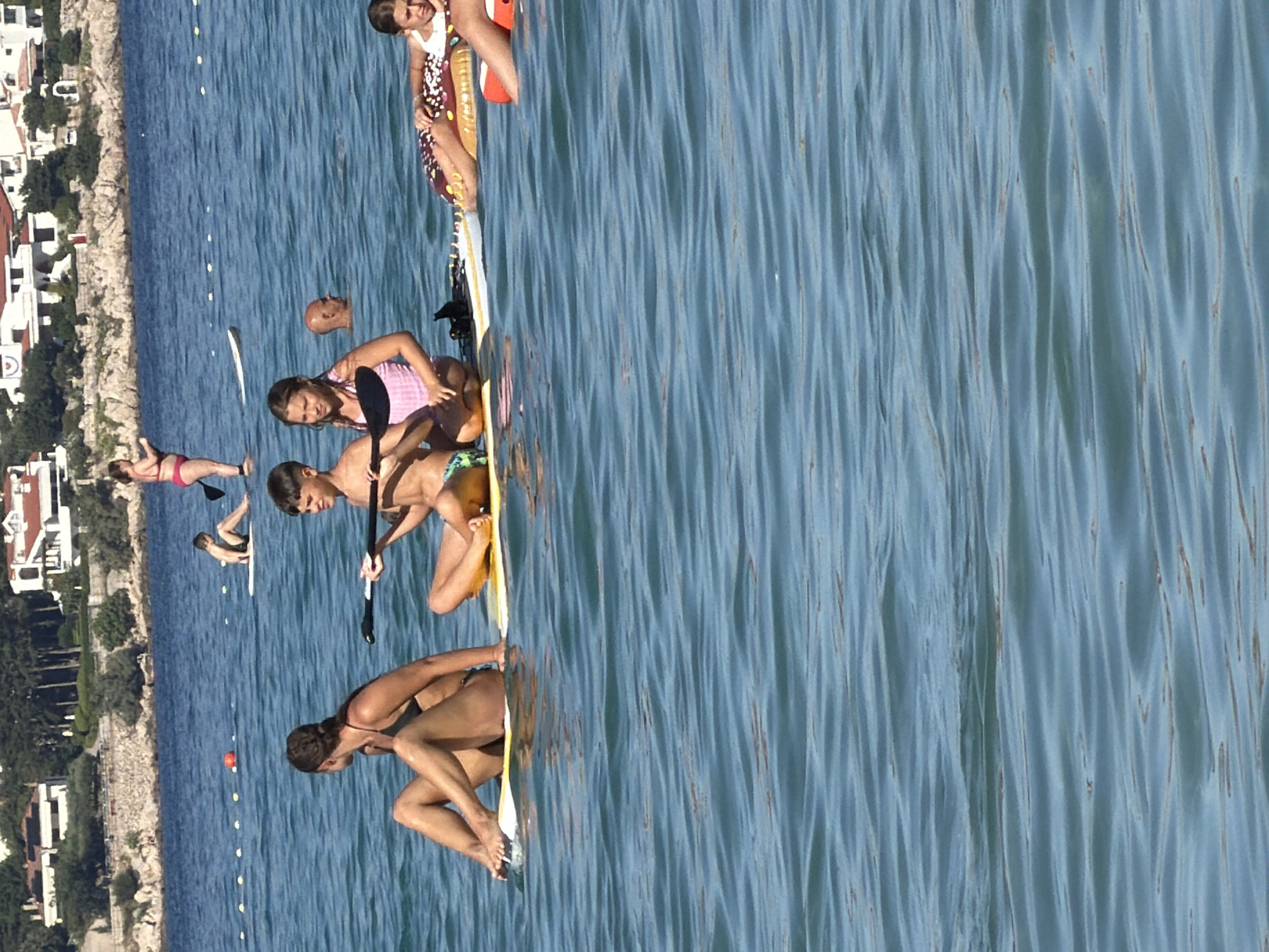 In Općina Baška, Croatia, on a clear, sunny day, several people are enjoying recreational activities on the water. In the foreground, a woman with dark hair tied back, wearing a black bikini, sits on a paddleboard, facing left. To her right, a boy with dark hair, wearing swimming trunks, is seated on another paddleboard, holding a paddle. To his right, a girl with blonde hair, wearing a pink and white striped swimsuit, is also seated on a paddleboard. Further to the right, another girl, wearing a white swimsuit, is lying on a inflatable donut-shaped float. In the background, a bald man is visible in the water, with only his head and shoulders showing. Another woman in a pink bikini stands on a paddleboard in the distance. The water is a deep blue, with visible ripples, and the coastline with white buildings and green trees is visible in the background.