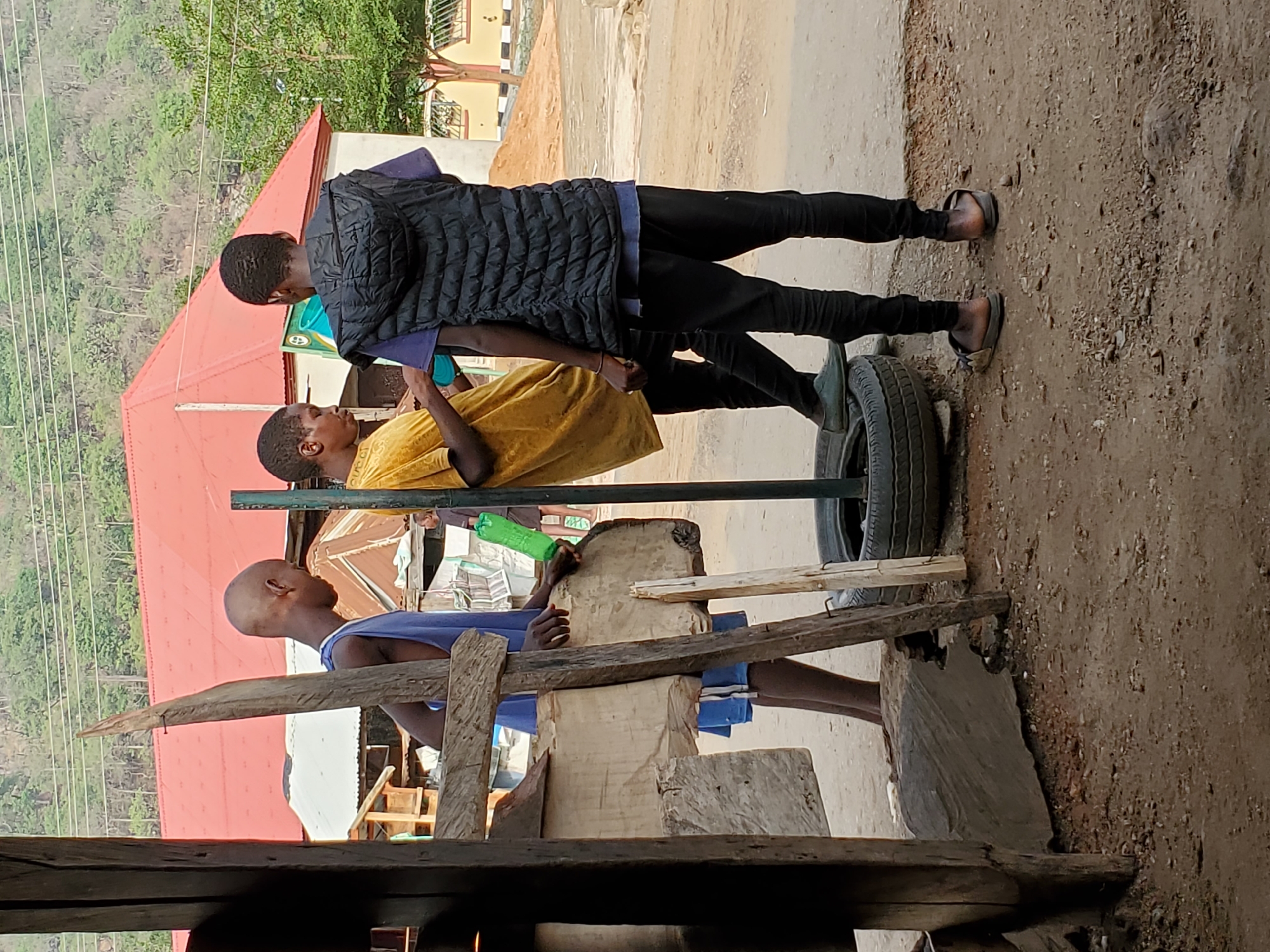 In Mararaba, Nigeria, a group of three young boys is outdoors. One boy in a blue tank top stands behind a wooden structure made of rough-cut lumber. Another boy, wearing a yellow t-shirt, is positioned behind a metal bar supported by a tire. A third boy stands behind the second, wearing a black vest. The setting appears to be a dirt or gravel surface adjacent to a building with a red roof. The background includes greenery and possibly other buildings.