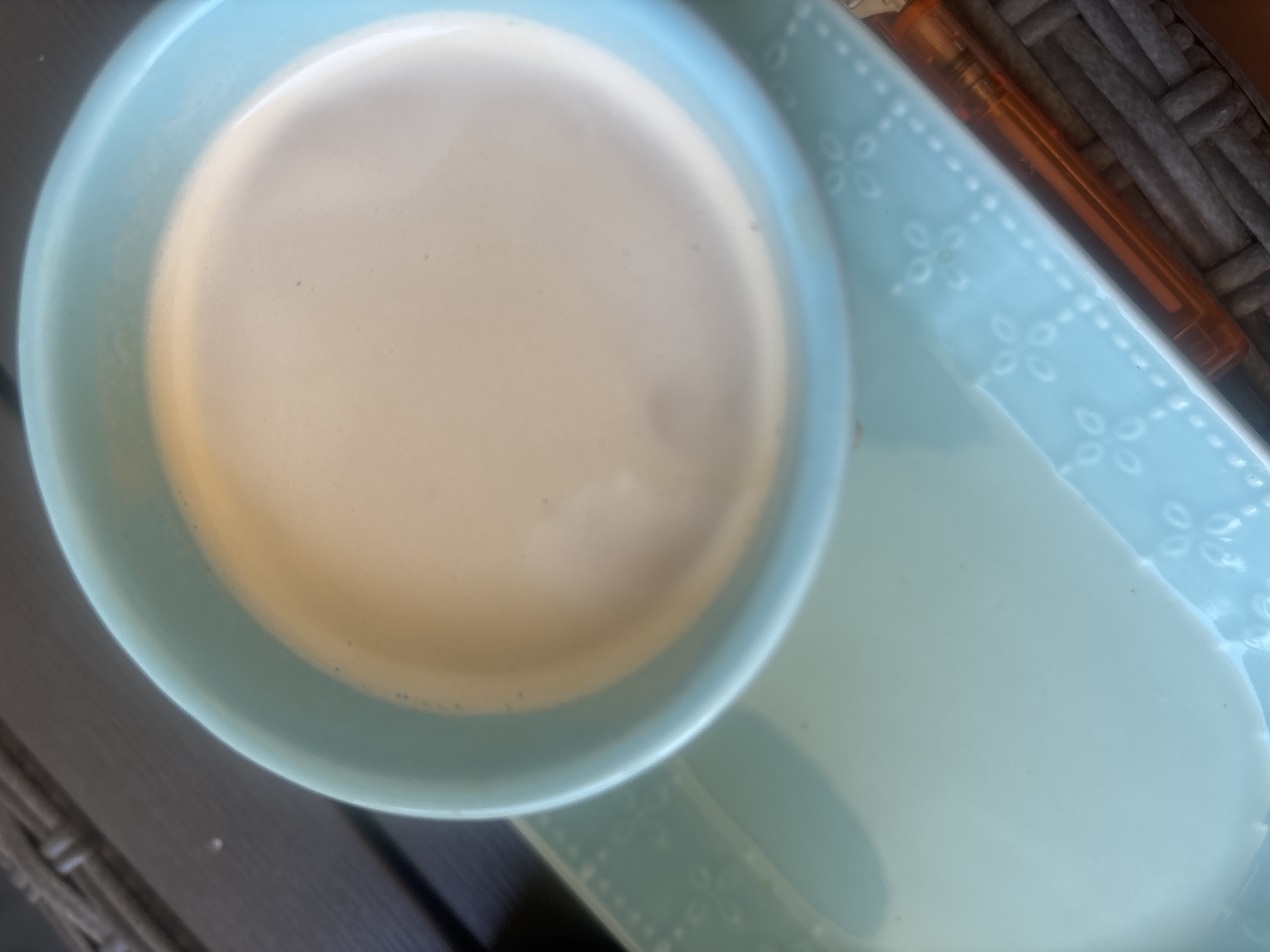 A close-up, top-down view captures a light blue ceramic bowl filled with a light brown, frothy beverage. The bowl rests on a matching light blue ceramic platter with a delicate embossed floral pattern. To the right of the platter, a bright orange object, possibly a pen or lighter, is visible, resting on a dark gray, woven surface. The background is blurred, suggesting an outdoor setting, but no specific Plovdiv landmarks or environmental features are identifiable.